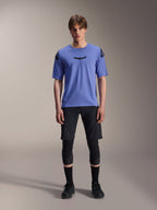 Mannelijk model draagt Alpinestars A-Aria Polartec Elite Jersey met korte mouwen in Baja Blue, zwart astars-logo op de borst, gecombineerd met zwarte MTB-short, kniebeschermers en zwarte fietsschoenen, studio-opname voor mountainbikekleding