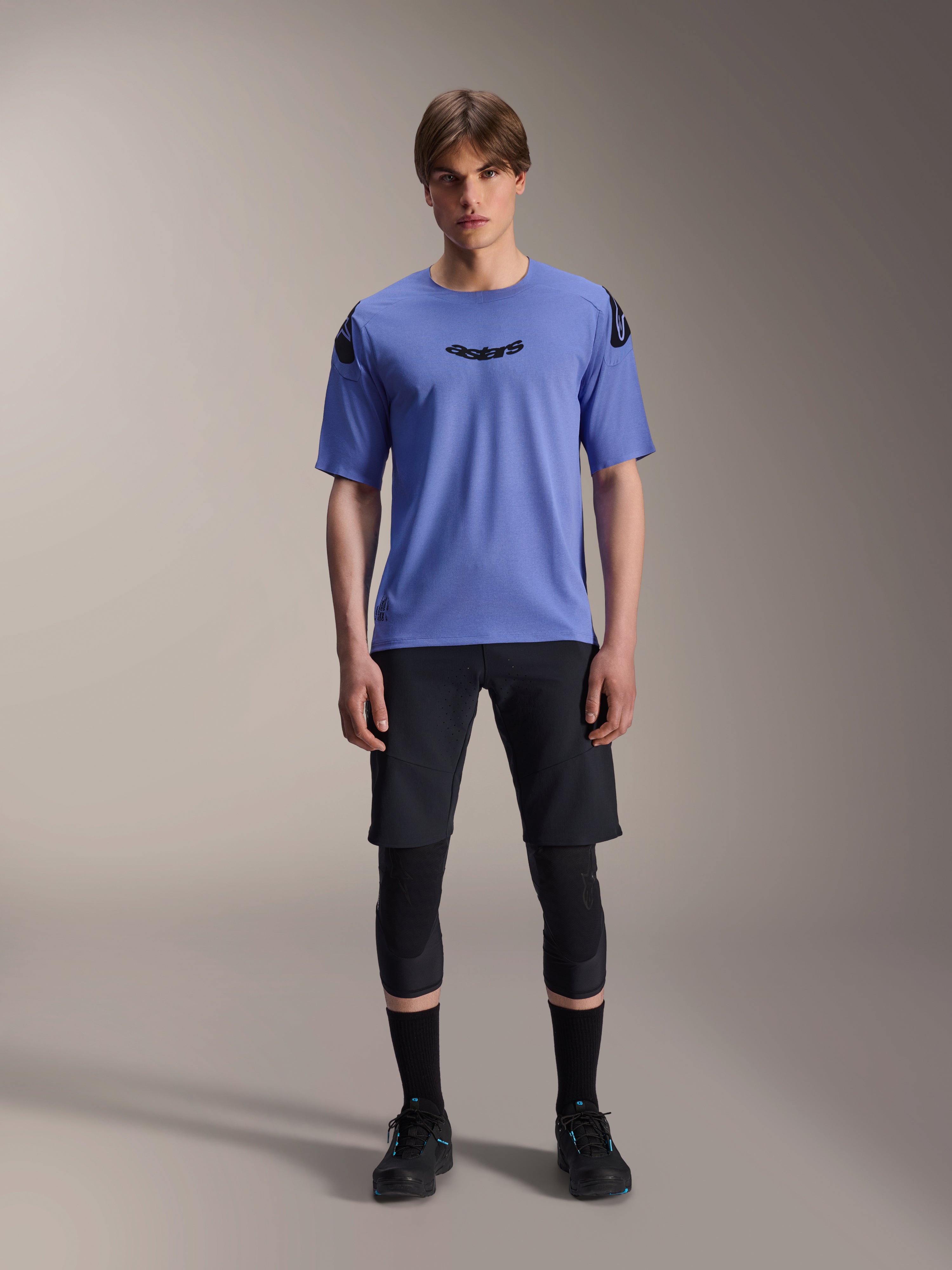 Mannelijk model draagt Alpinestars A-Aria Polartec Elite Jersey met korte mouwen in Baja Blue, zwart astars-logo op de borst, gecombineerd met zwarte MTB-short, kniebeschermers en zwarte fietsschoenen, studio-opname voor mountainbikekleding