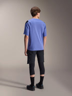 Alpinestars A-Aria Polartec Elite Jersey met korte mouwen in Baja Blue, achteraanzicht op een mannelijk model, gecombineerd met een zwarte MTB-short en kniebescherming, voorzien van een performance-pasvorm en verlengde achterkant voor trailrijden.