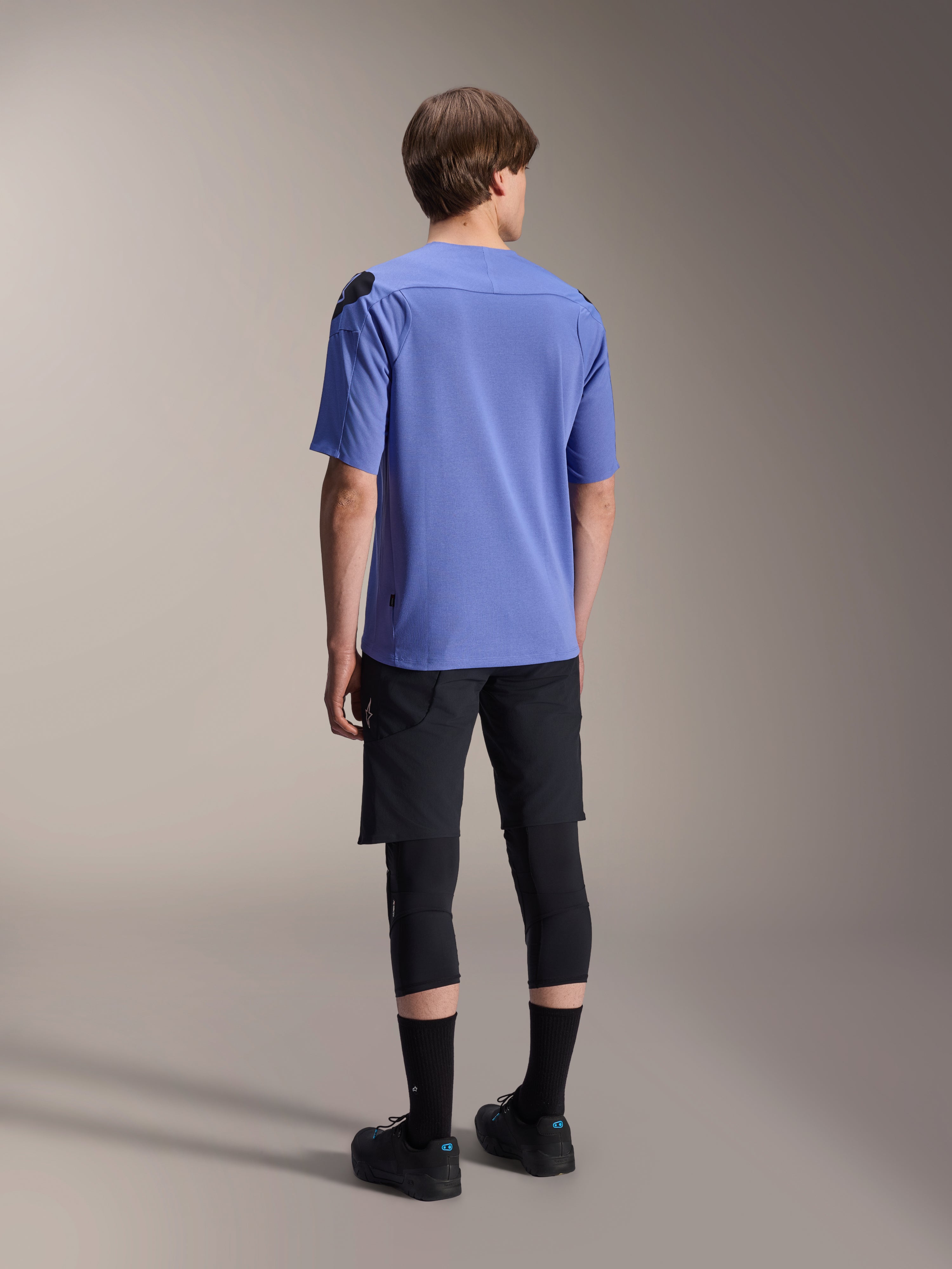 Alpinestars A-Aria Elite shorts, mountainbike short, Black Titanium, achteraanzicht gedragen met een lavendelkleurige jersey met korte mouwen, zwarte kniebeschermers en zwarte fietsschoenen, sportieve MTB-uitrusting.
