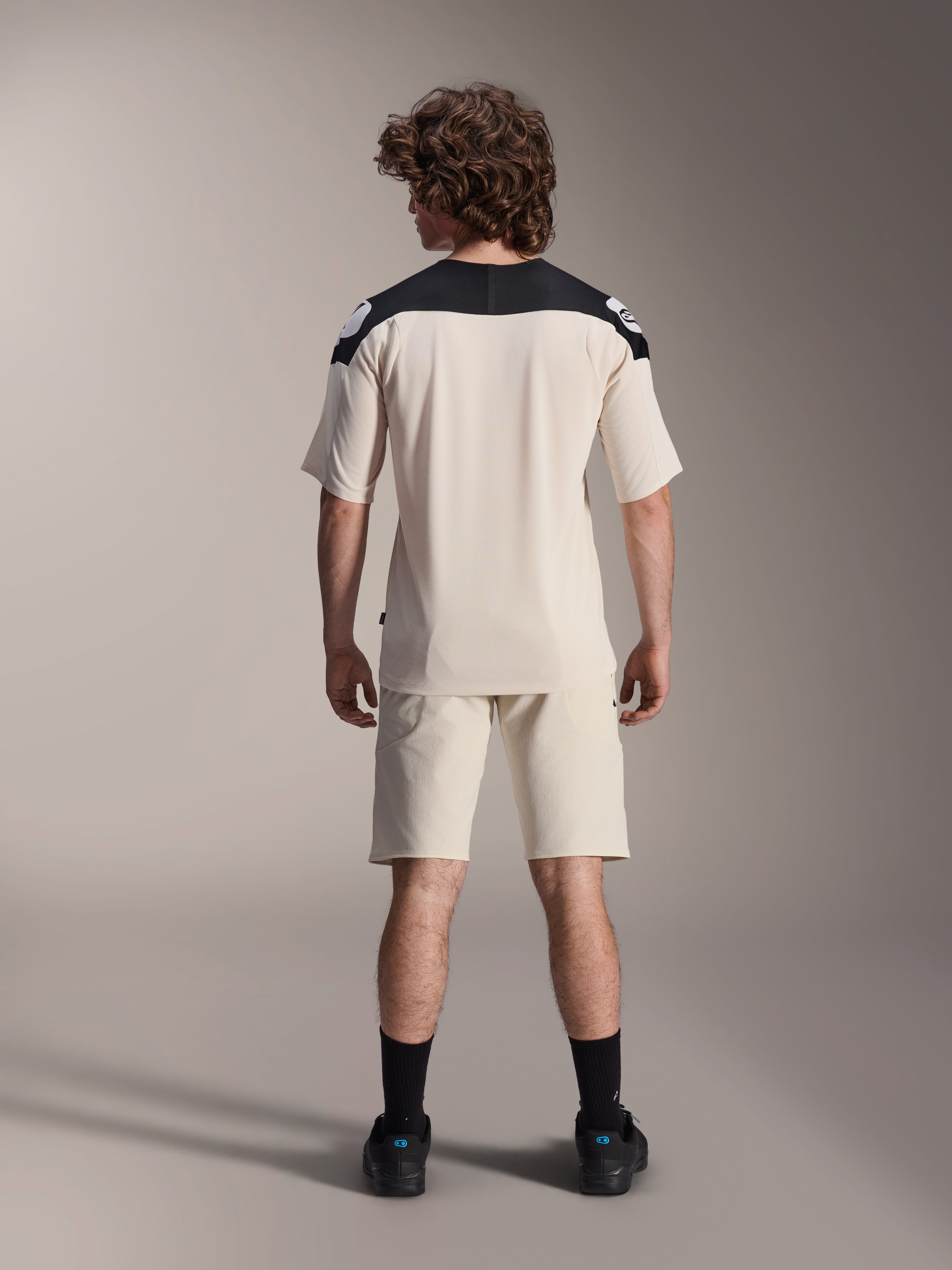 Alpinestars A-Aria Polartec Elite Jersey met korte mouwen, MTB-fietsshirt, ecru met zwarte schouderpanelen, achteraanzicht getoond met bijpassende short en zwarte sokken, prestatiegerichte pasvorm met een verlengde achterkant voor off-road rijden.