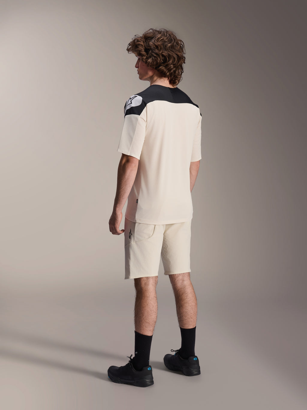 Alpinestars A-Aria Elite shorts en bijpassend shirt, mountainbikekleding, ecru met zwarte accenten, mannelijk model van achteren gezien met zwarte sokken en schoenen, performance off-road fietskleding