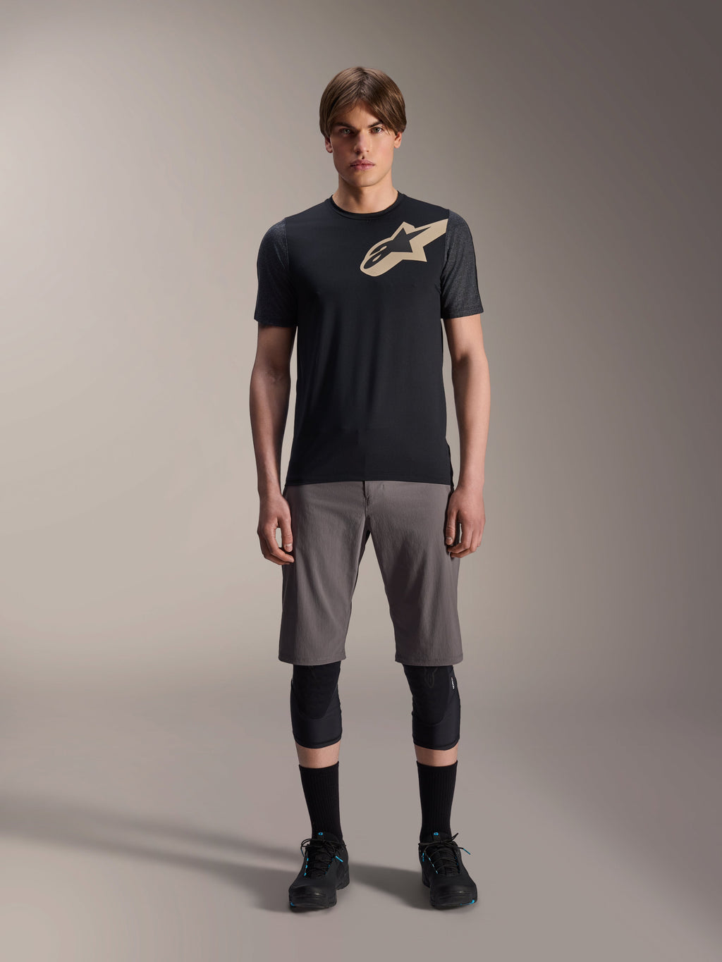 Alpinestars A-Dura Lipan Jersey met korte mouwen, zwart met grijs gemêleerde mouwen en zandkleurig Astars-logo op de borst, gedragen door een mannelijk model met een grijze mountainbikeshort en zwarte kniebeschermers, technische MTB-kleding