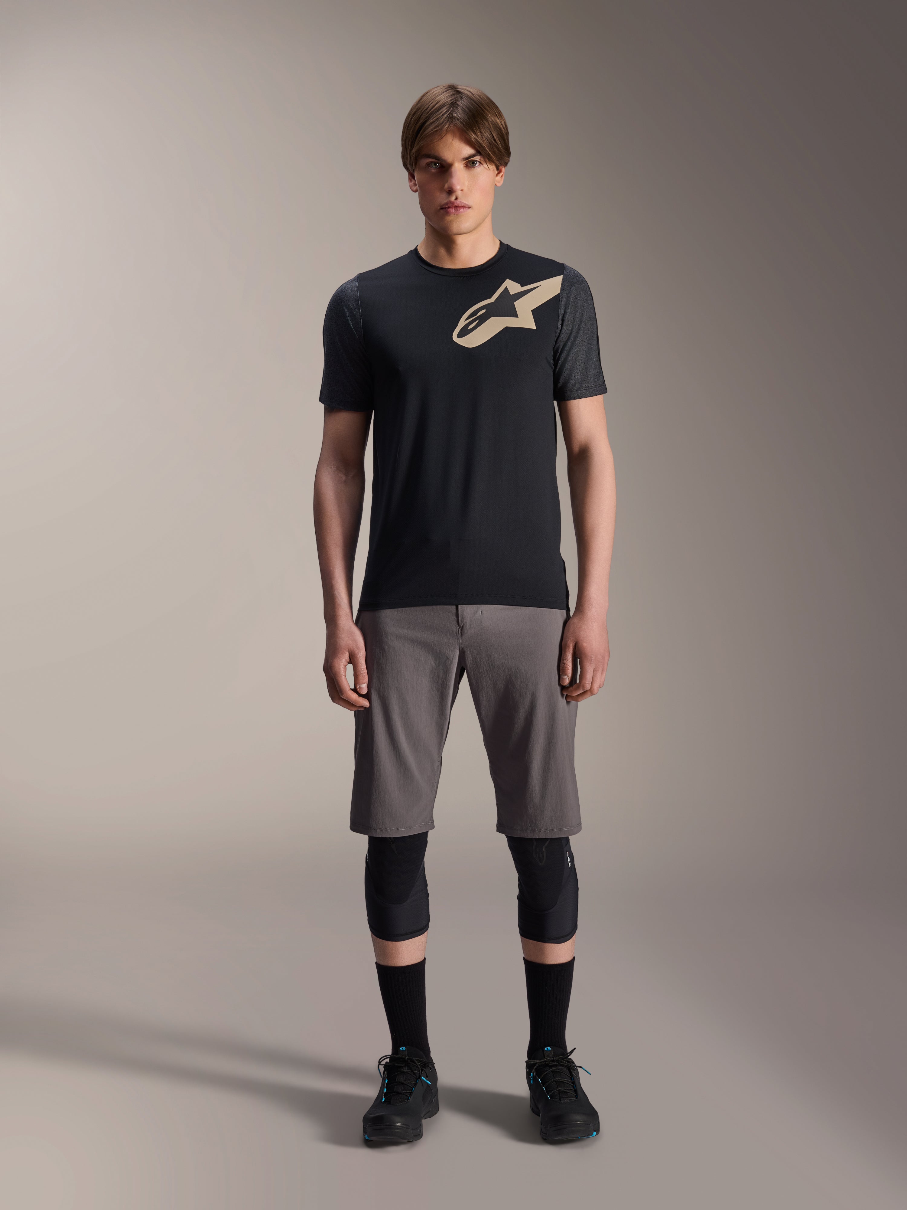 Alpinestars A-Dura Lipan Jersey met korte mouwen, zwart met grijs gemêleerde mouwen en zandkleurig Astars-logo op de borst, gedragen door een mannelijk model met een grijze mountainbikeshort en zwarte kniebeschermers, technische MTB-kleding