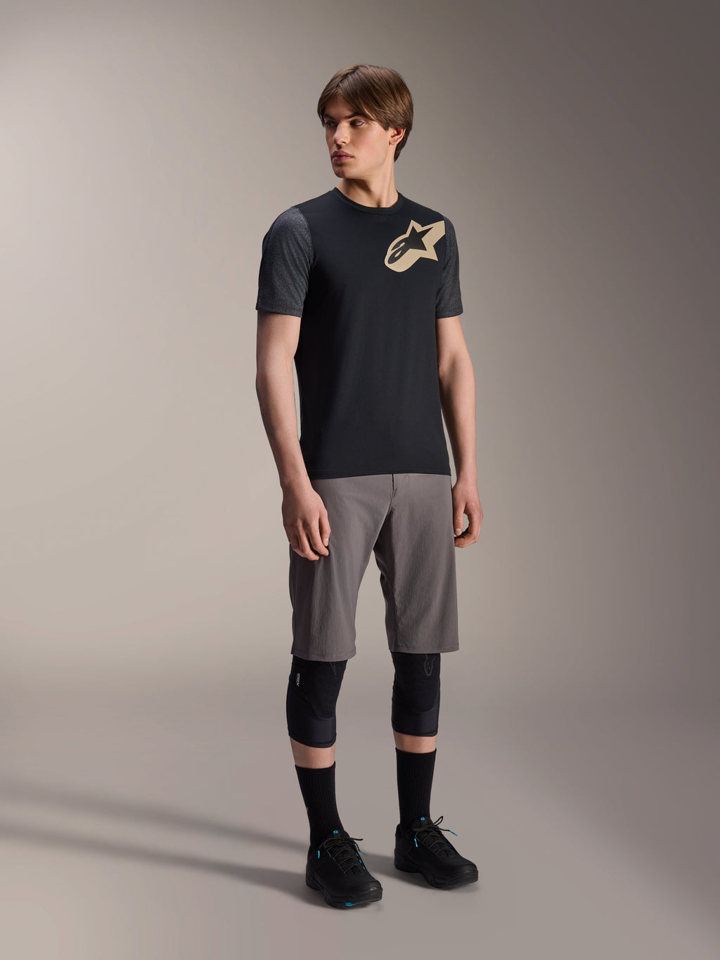 Mannelijk model in Alpinestars A-Dura Lipan jersey met korte mouwen in het zwart, met donkergrijze mouwen en een beige Astars-logo op de borst, gecombineerd met een grijze mountainbikeshort, zwarte kniebeschermers en zwarte fietsschoenen, op elkaar afgestemde mountainbike-uitrusting