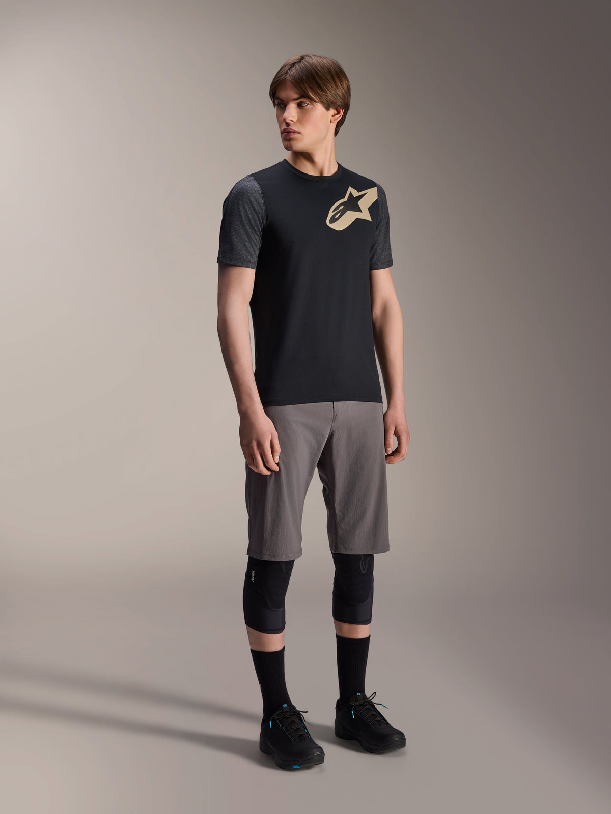 Mannelijk model in Alpinestars A-Dura Lipan jersey met korte mouwen in het zwart, met donkergrijze mouwen en een beige Astars-logo op de borst, gecombineerd met een grijze mountainbikeshort, zwarte kniebeschermers en zwarte fietsschoenen, op elkaar afgestemde mountainbike-uitrusting