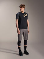 Mannelijk model in Alpinestars A-Dura Lipan jersey met korte mouwen in het zwart, met donkergrijze mouwen en een beige Astars-logo op de borst, gecombineerd met een grijze mountainbikeshort, zwarte kniebeschermers en zwarte fietsschoenen, op elkaar afgestemde mountainbike-uitrusting