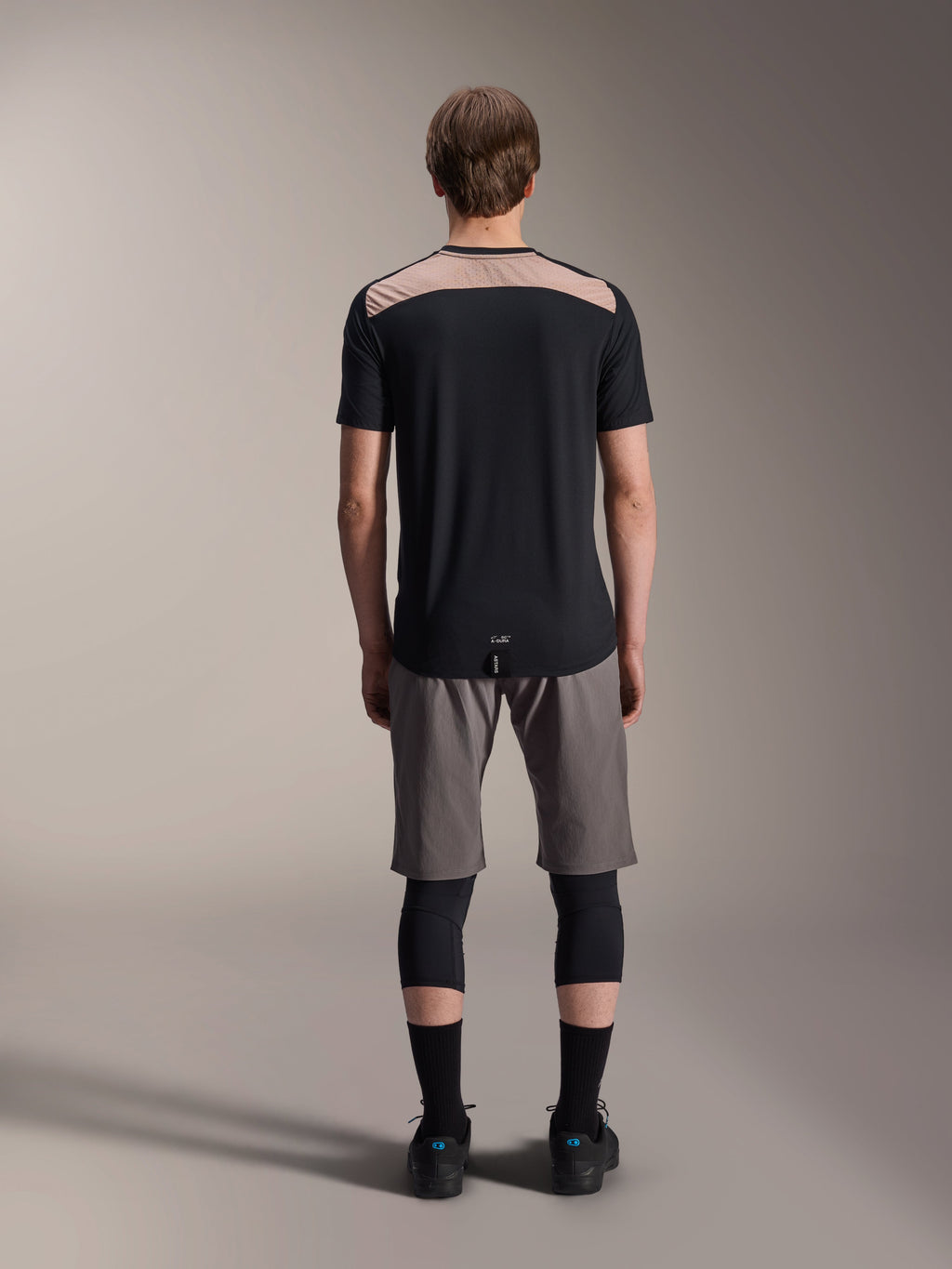 Alpinestars A-Dura Lipan Jersey - Korte mouwen, zwart met tan bovenpaneel op de rug, achteraanzicht met verlengde achterkant, gedragen met grijze MTB-short en zwarte kniebeschermers, technische mountainbikekleding