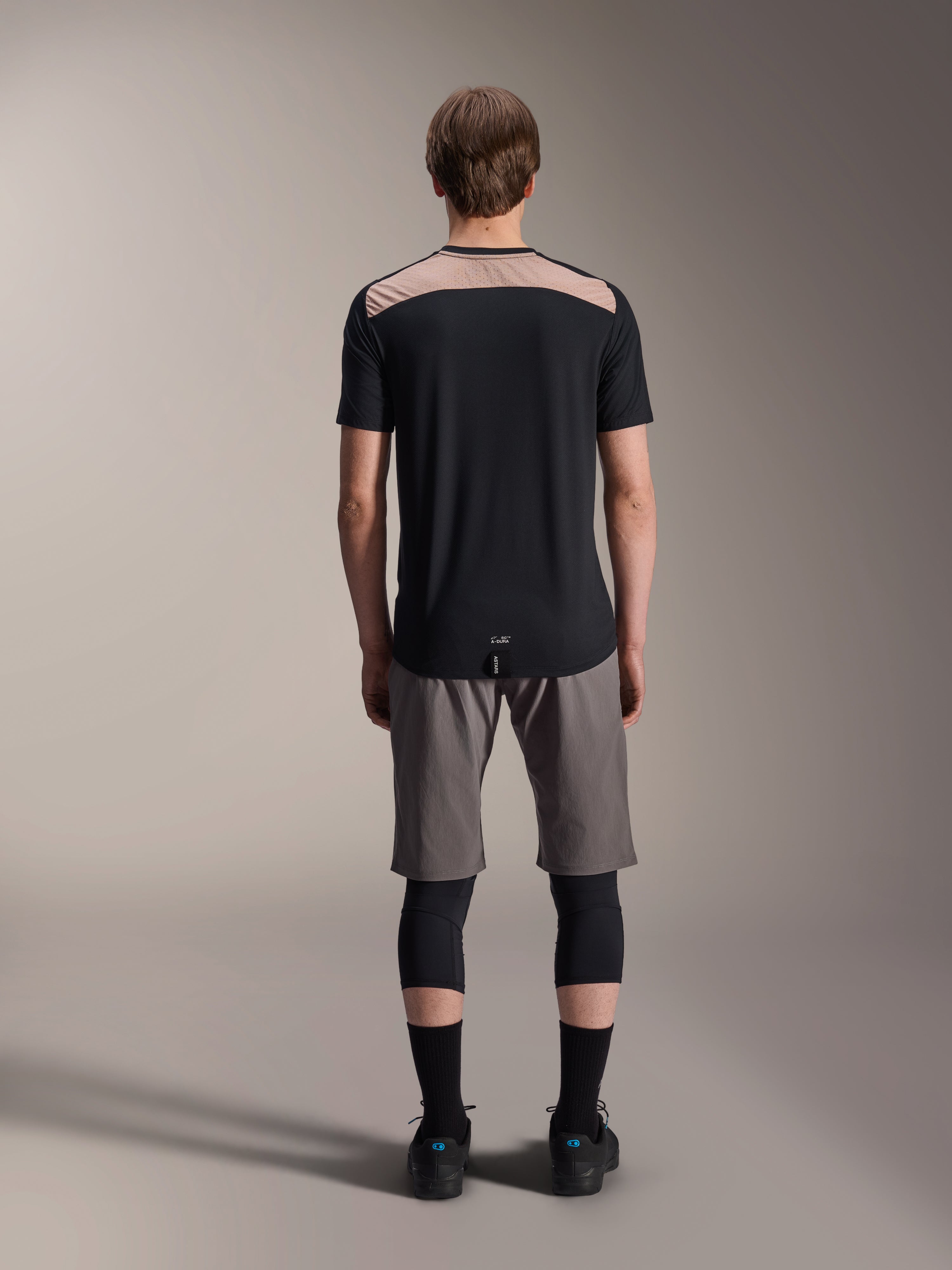 Alpinestars A-Dura Lipan Jersey - Korte mouwen, zwart met tan bovenpaneel op de rug, achteraanzicht met verlengde achterkant, gedragen met grijze MTB-short en zwarte kniebeschermers, technische mountainbikekleding