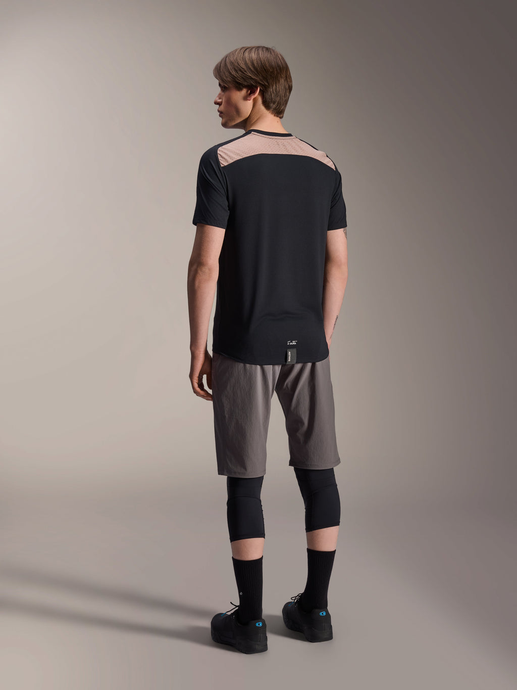 Alpinestars A-Dura Lipan Jersey met korte mouwen in het zwart, achteraanzicht op model met het lichtbruine geperforeerde mesh-paneel op de bovenrug, gecombineerd met een grijze mountainbikebroek en zwarte kniebeschermers voor een complete trail-rijoutfit.