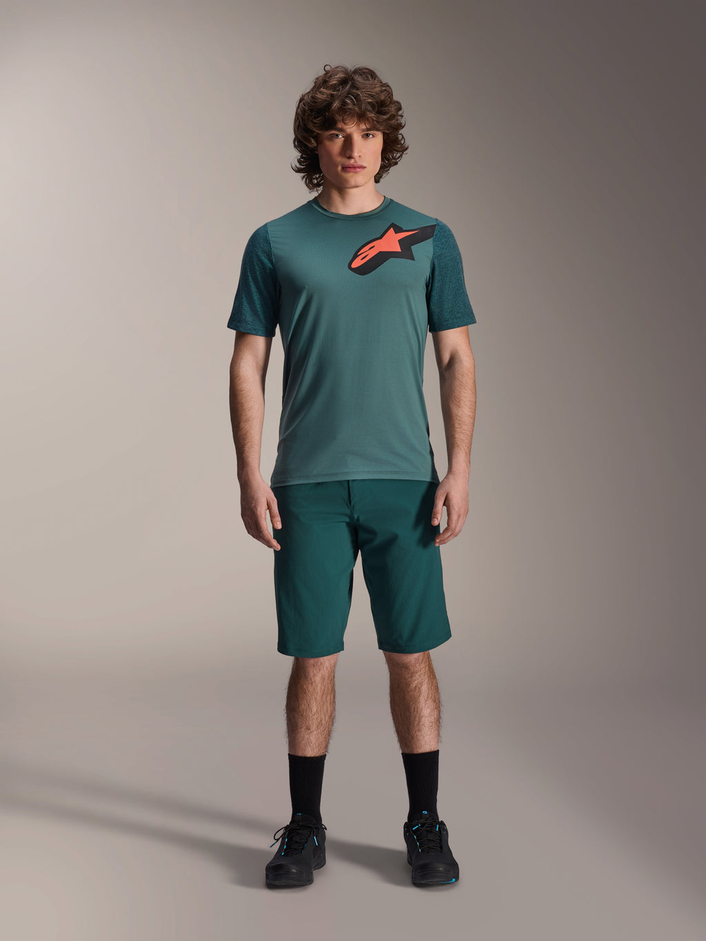 Alpinestars A-Dura Lipan Jersey, MTB-shirt met korte mouwen, Silver Pine groen met zwart en oranje Astars-logo op de borst, gecombineerd met bijpassende shorts en zwarte schoenen, technische mountainbikekleding