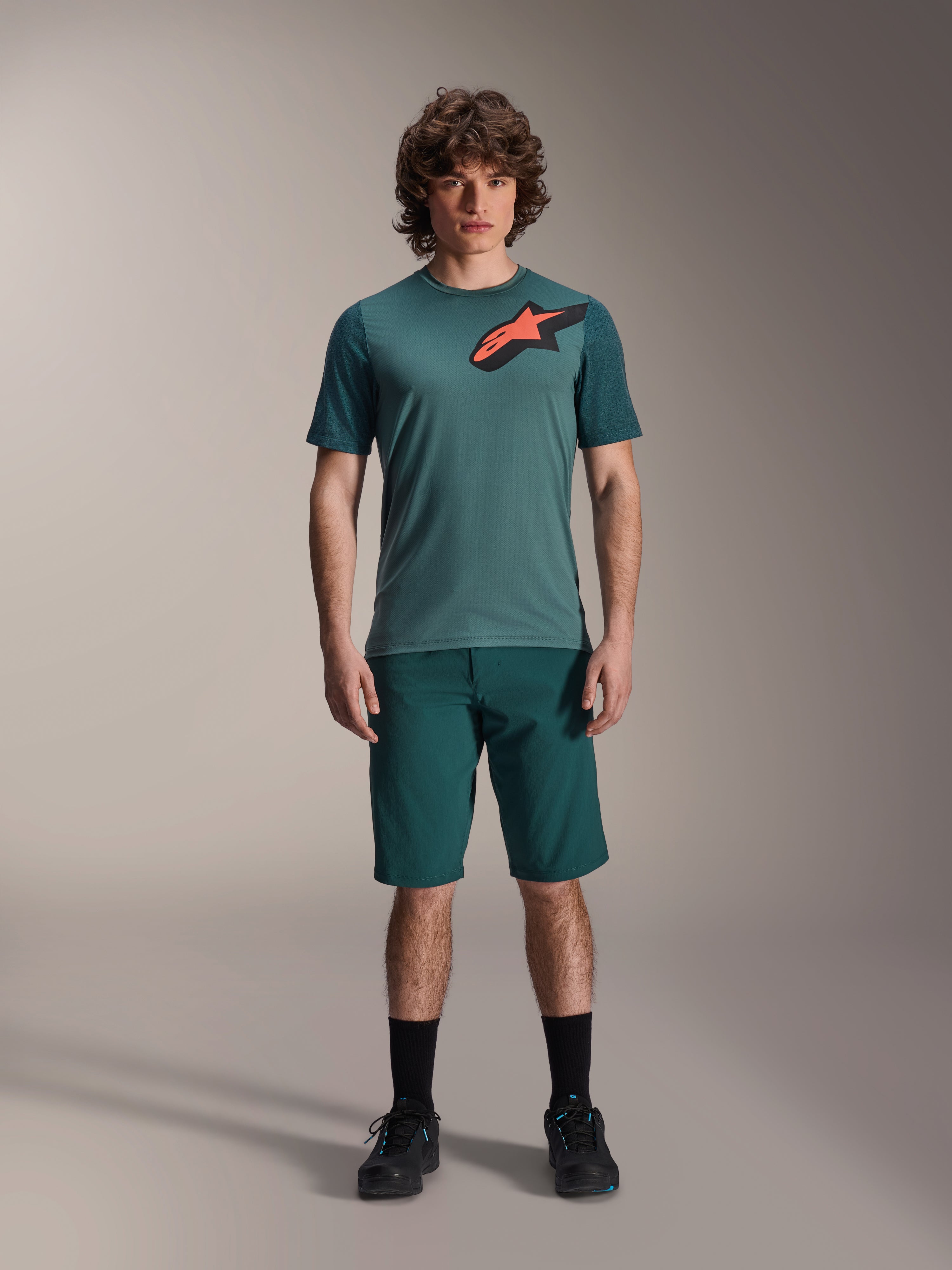 Alpinestars A-Dura Shorts in Dark Pine, mountainbike-short voor heren, getoond op een model met een bijpassende petrolkleurige jersey met korte mouwen met een rood en zwart sterlogo, zwarte sokken en fietsschoenen, casual sportieve mountainbike-kleding