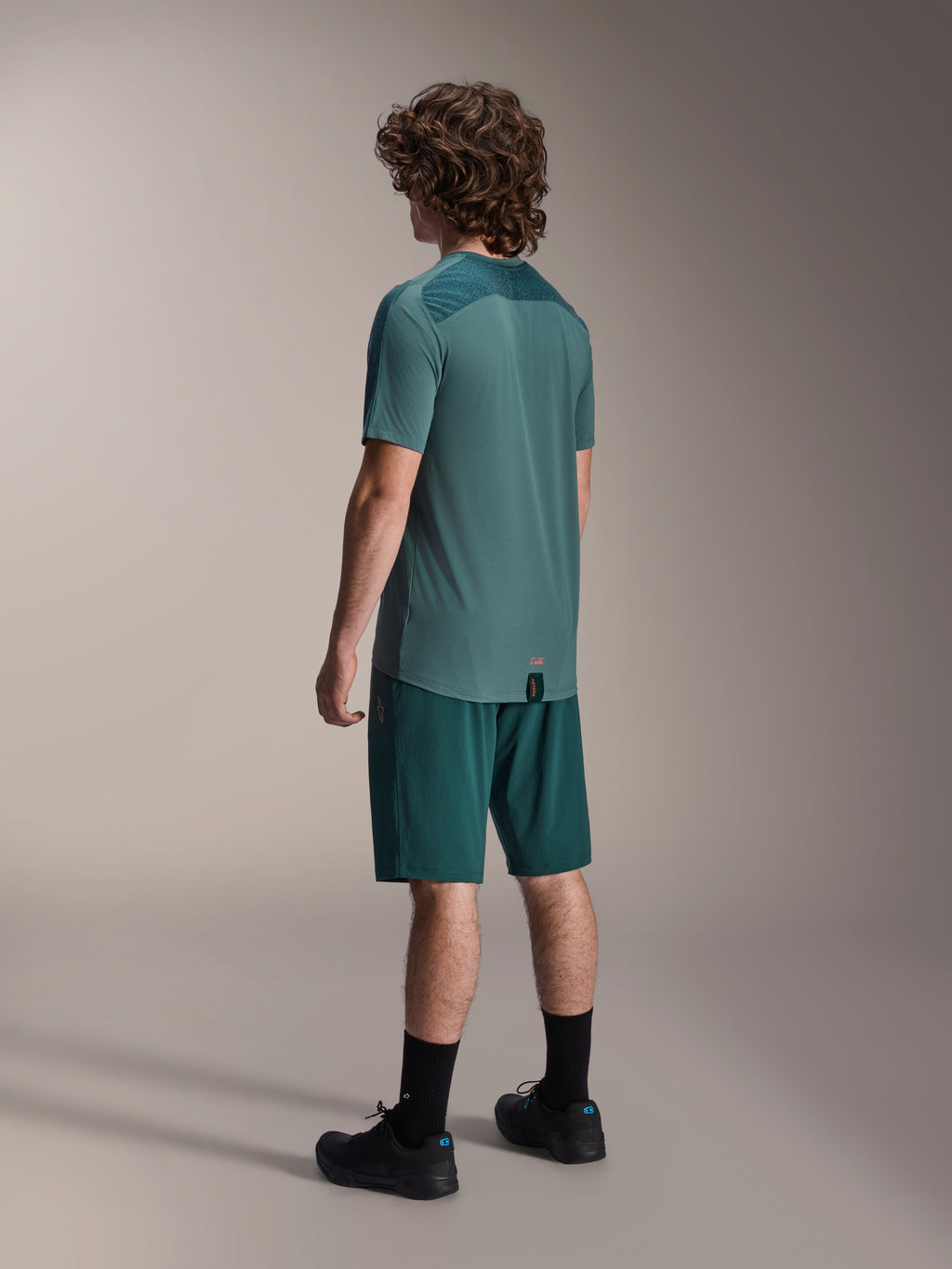 Model draagt Alpinestars A-Dura Shorts in Dark Pine en een bijpassende groene Lipan jersey met korte mouwen, achteraanzicht, gecombineerd met zwarte sokken en zwarte mountainbikeschoenen, professionele MTB-rijuitrusting.
