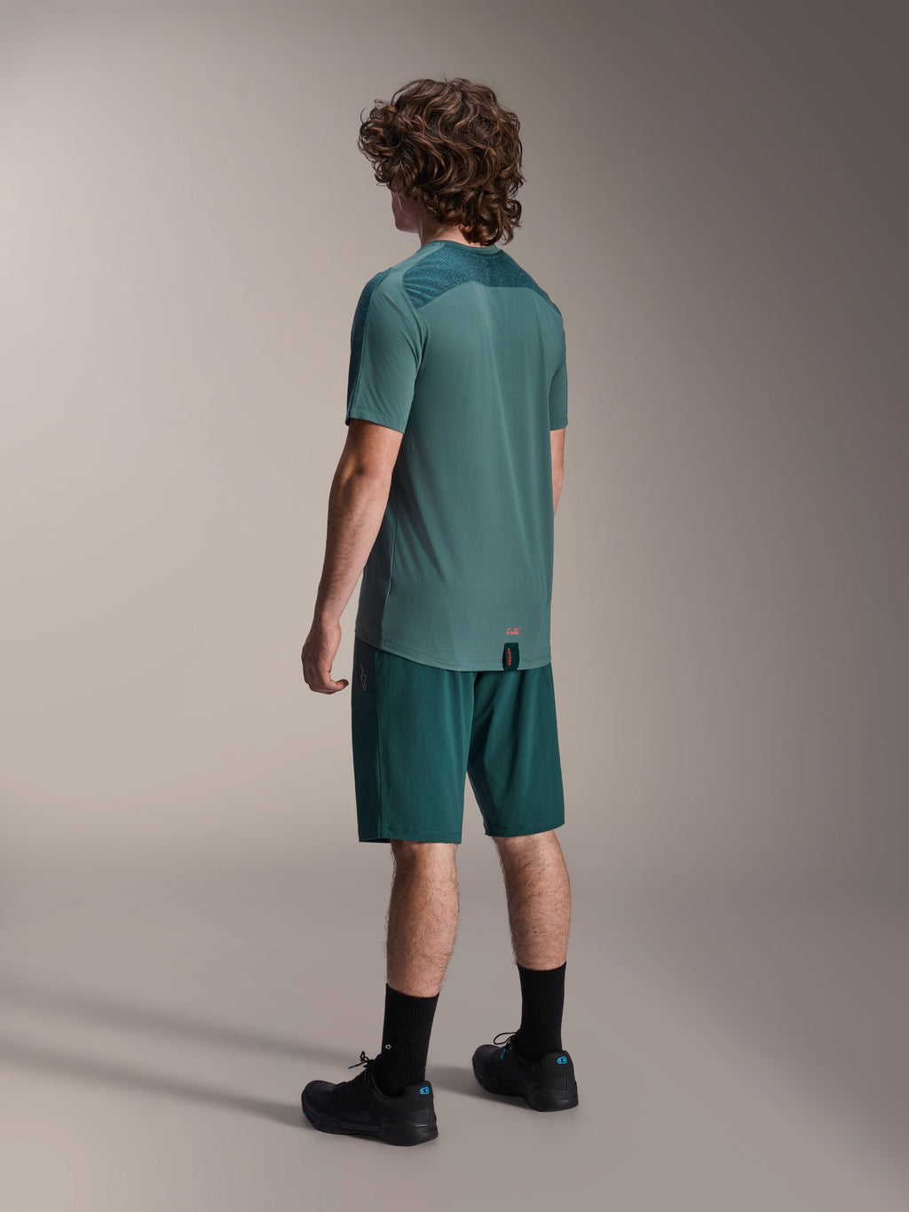 Alpinestars A-Dura Lipan jersey met korte mouwen in Silver Pine, achteraanzicht van een mannelijk model met bijpassende donkergroene mountainbike shorts, zwarte sokken en schoenen, voorzien van een getextureerd rugpaneel en een verlengde achterzoom voor extra bedekking.