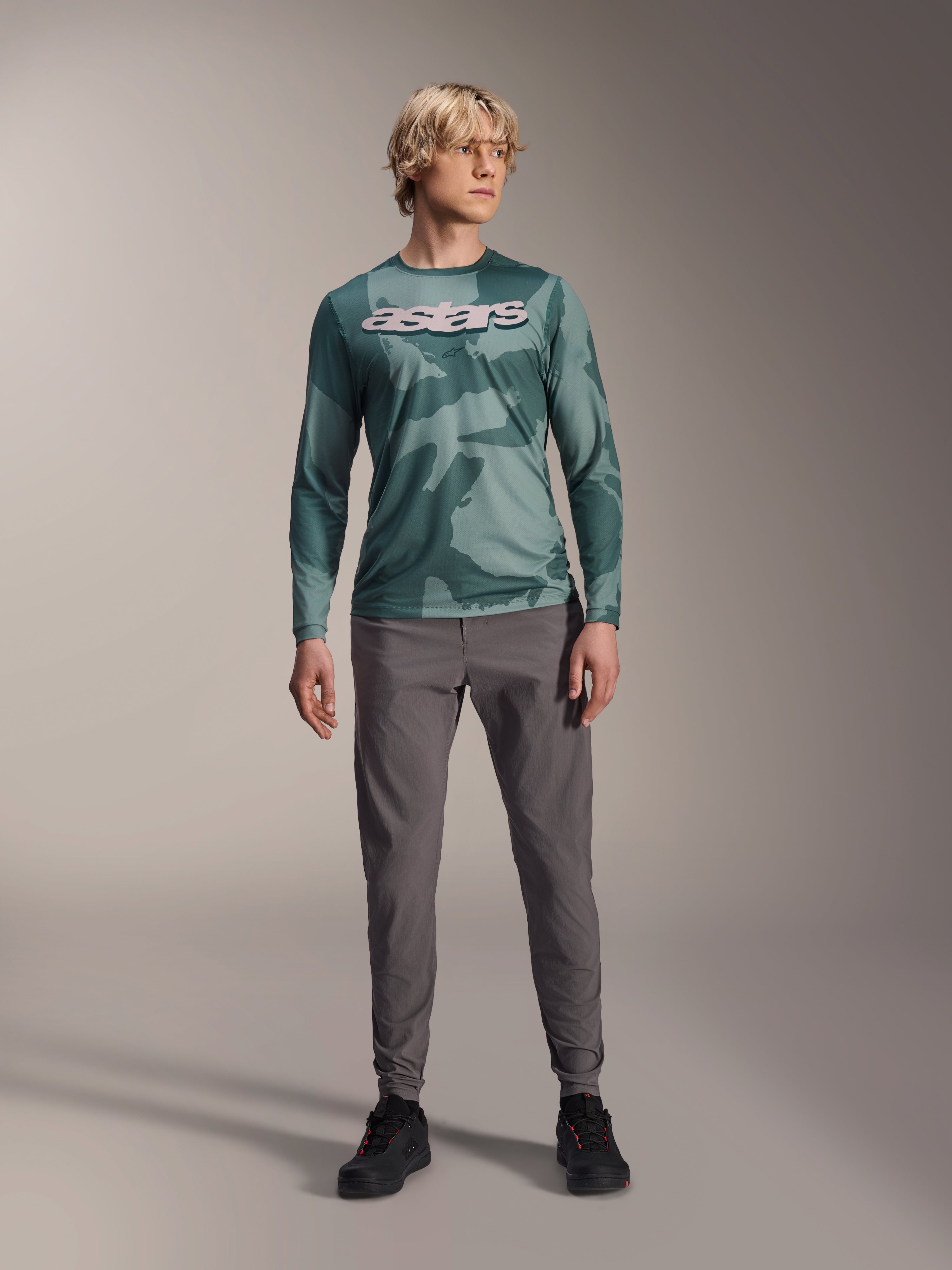 Alpinestars A-Dura Hollow Camo jersey met lange mouwen, Silver Pine Camo met groen camouflagepatroon, groot wit "astars" logo op de borst, gedragen met grijze technische broek en zwarte fietsschoenen, hoogwaardige MTB-uitrusting