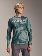 Alpinestars A-Dura Hollow Camo Jersey met lange mouwen, MTB-shirt, groen camouflagepatroon in Silver Pine Camo, groot lichtgrijs astars-logo op de borst, gedragen met een grijze broek, hoogwaardige mountainbikekleding