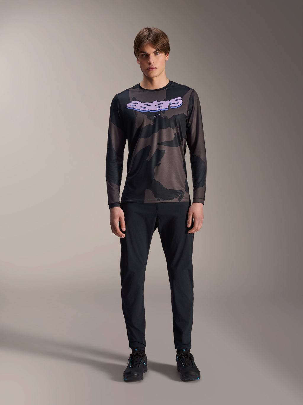 Model draagt Alpinestars A-Dura Hollow Camo Jersey met lange mouwen in Black Camo, voorzien van een paars 'astars' logo op de borst, gecombineerd met een zwarte broek en zwarte fietsschoenen, mountainbike-kleding