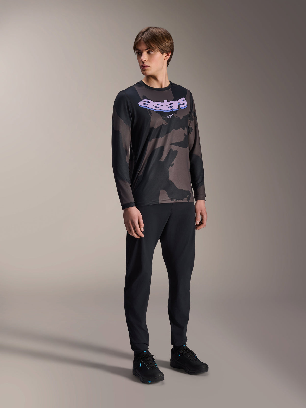Mannelijk model draagt de Alpinestars A-Dura Hollow Camo Jersey met lange mouwen, Black Camo-patroon met paarse 'astars' grafische opdruk op de borst, gecombineerd met een zwarte technische broek en zwarte schoenen, casual MTB-uitrusting