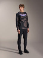 Mannelijk model draagt de Alpinestars A-Dura Hollow Camo Jersey met lange mouwen, Black Camo-patroon met paarse 'astars' grafische opdruk op de borst, gecombineerd met een zwarte technische broek en zwarte schoenen, casual MTB-uitrusting
