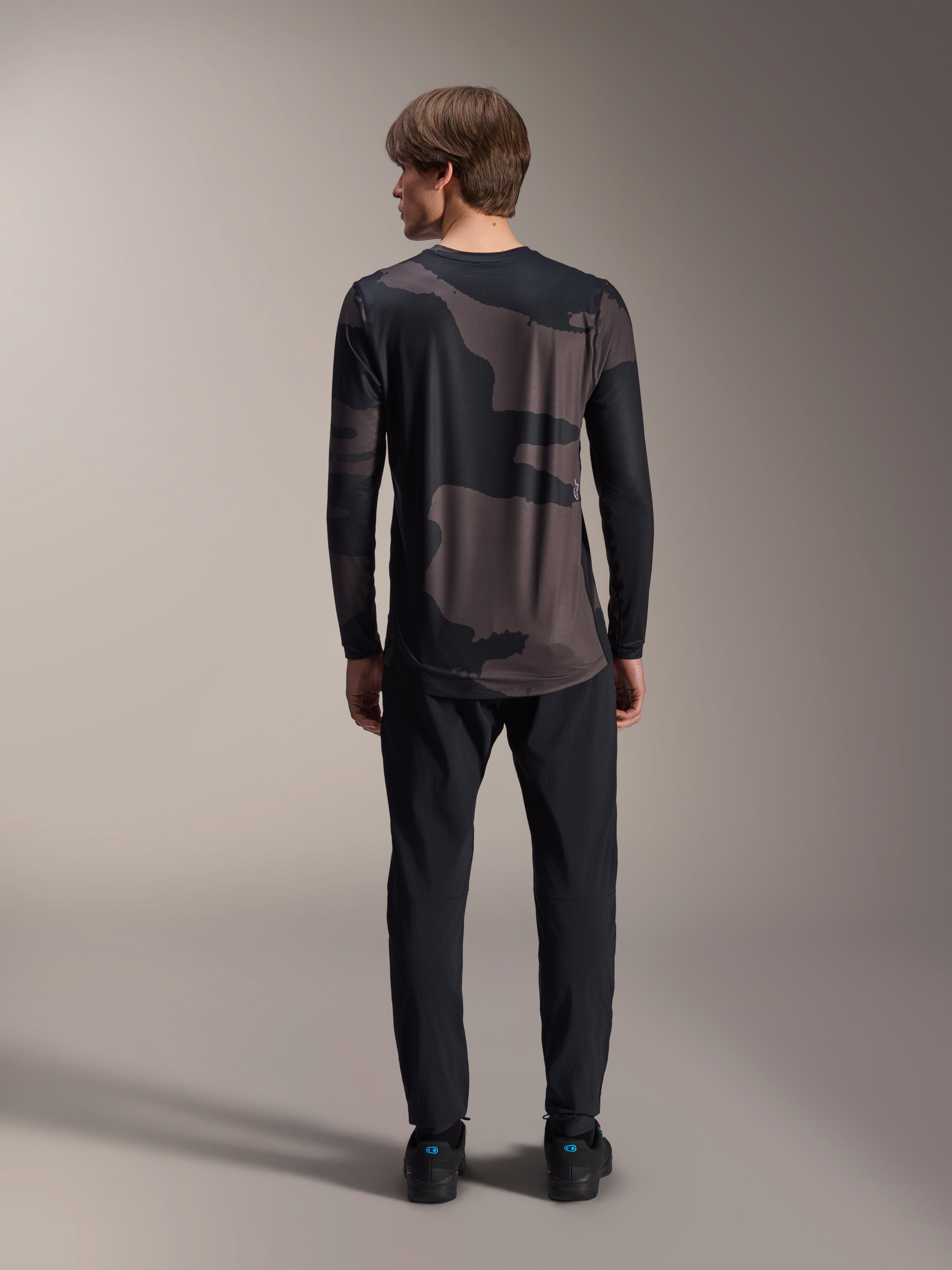 Alpinestars A-Dura Hollow Camo Jersey met lange mouwen, Black Camo, achteraanzicht gedragen met een zwarte broek, voorzien van een donker camouflagepatroon en een verlengde achterkant voor MTB-dekking.