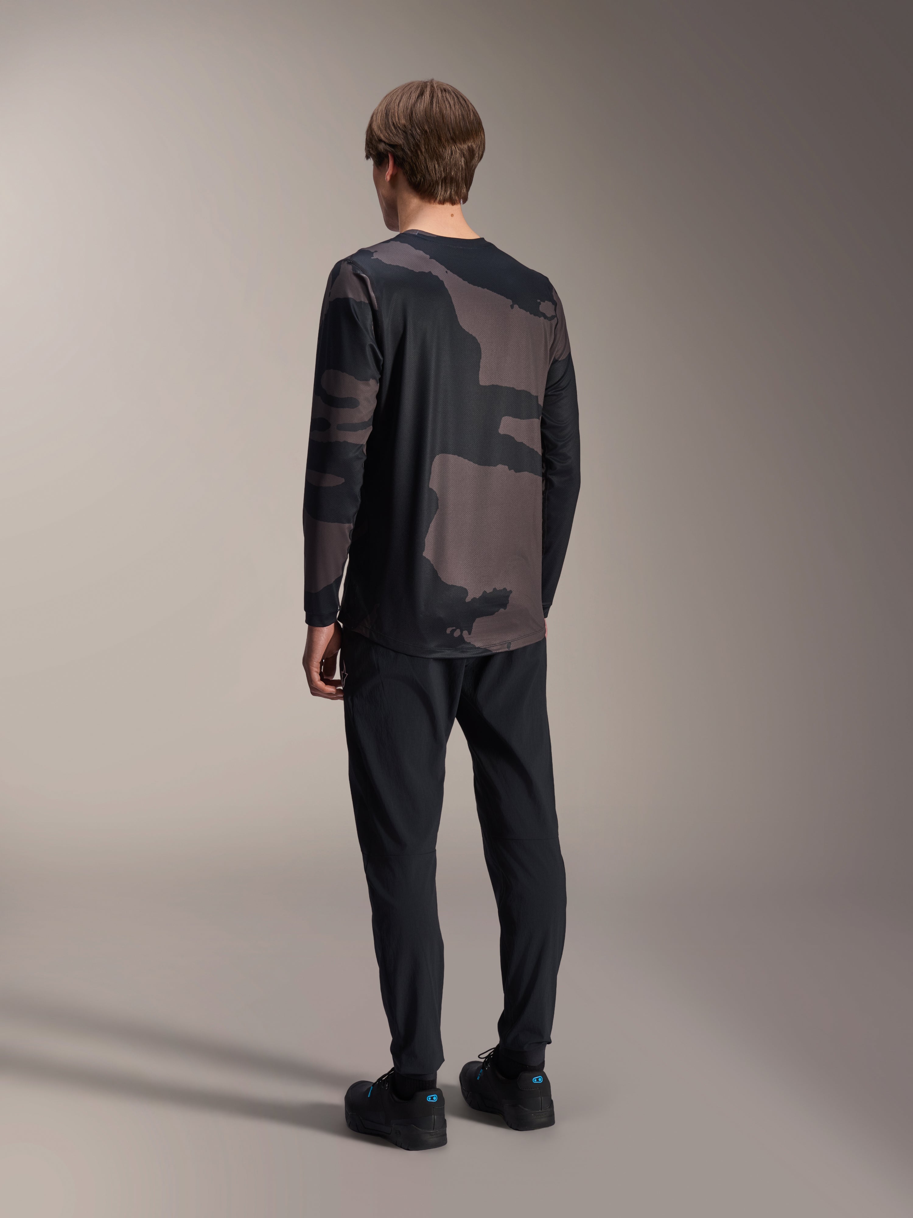 Alpinestars A-Dura Hollow Camo Jersey met lange mouwen, Black Camo, achteraanzicht van model, technisch mountainbikeshirt met verlengde achterkant, gecombineerd met een zwarte broek en fietsschoenen, casual off-road prestatiekleding