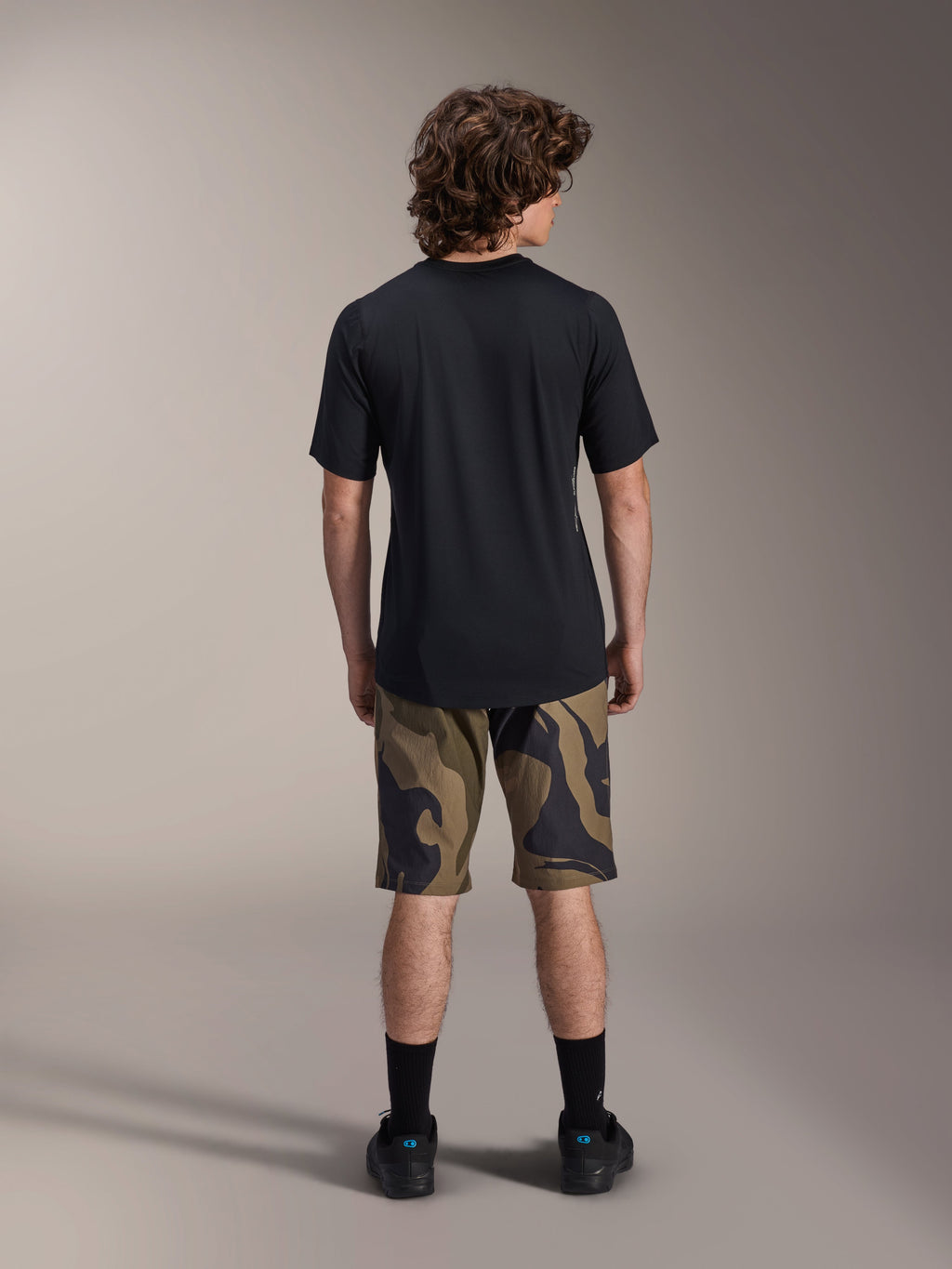 Alpinestars A-Dura Astar Jersey met korte mouwen, zwart, achteraanzicht op een model gecombineerd met MTB-shorts met camouflagepatroon en zwart schoeisel, casual technische mountainbikekleding