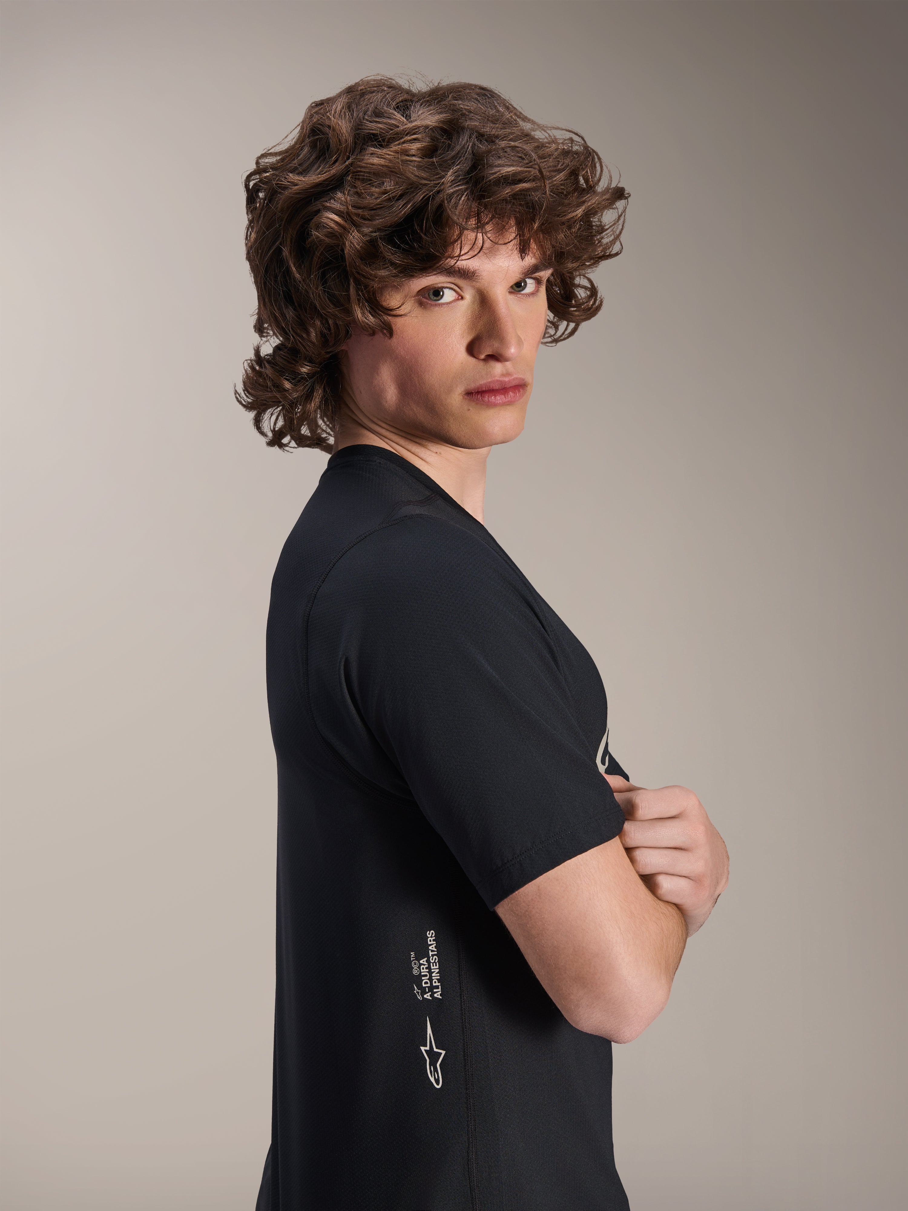 Alpinestars A-Dura Astar Jersey, MTB-shirt met korte mouwen, zwart, mannelijk model poserend in profiel met de armen over elkaar, voorzien van witte Alpinestars-branding aan de onderzijde, casual sportief ontwerp voor mountainbiken.