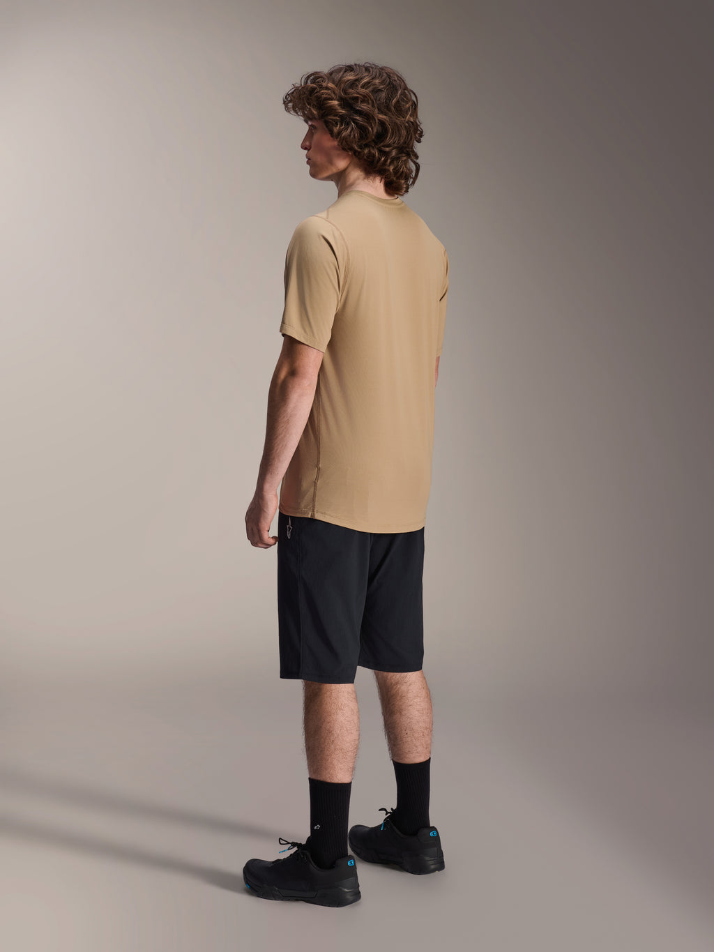 Alpinestars A-Dura Liner Shorts in zwart en A-Dura SS Jersey in tan, gedragen door een mannelijk model in zijaanzicht, gecombineerd met zwarte sokken en fietsschoenen, mountainbike lifestyle kleding.