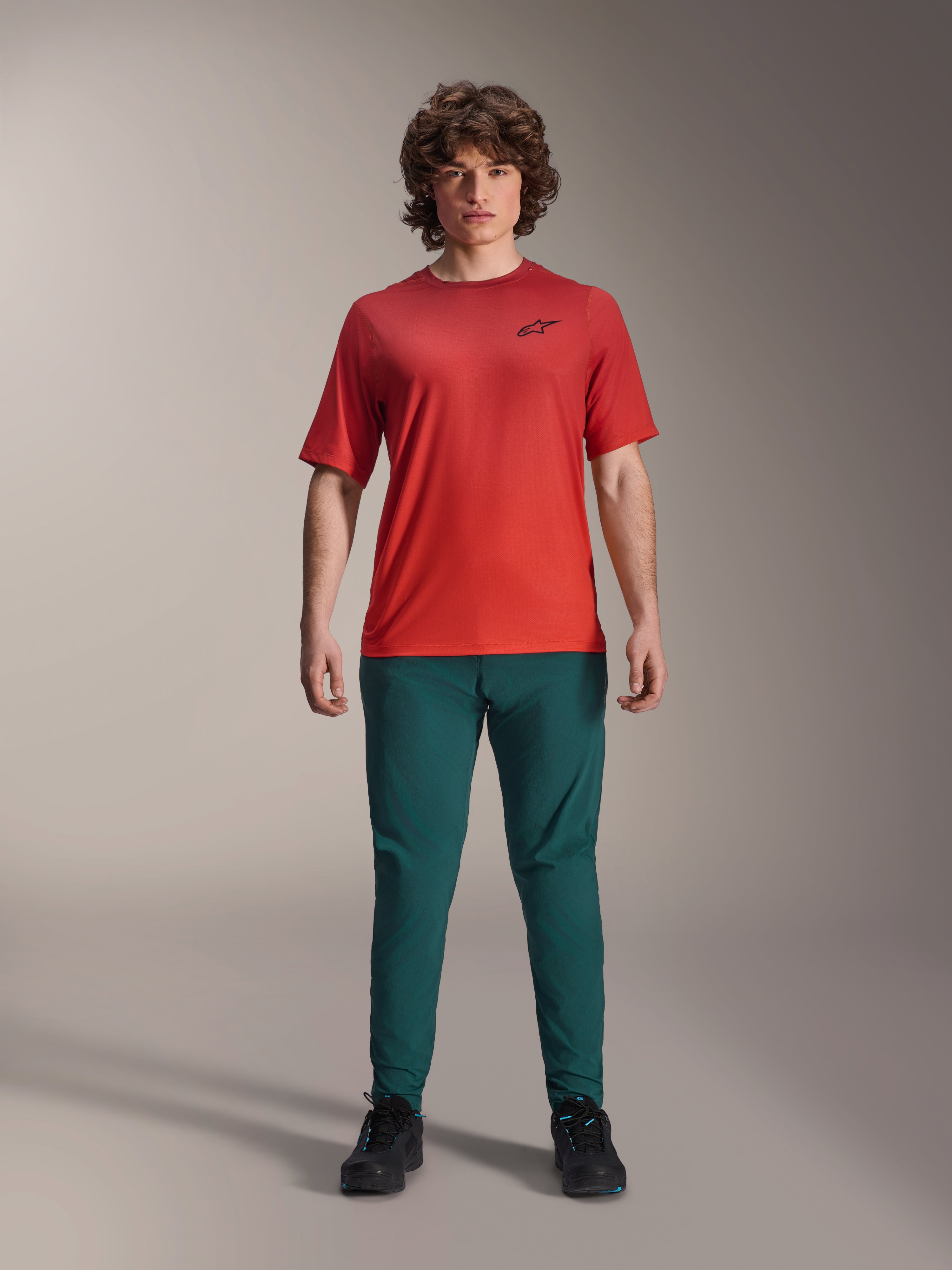 Alpinestars A-Dura Rise Jersey met korte mouwen, Spicy Orange, gedragen met een donkergroene broek en zwarte schoenen, voorzien van een zwart Astars-logo op de borst, casual performance mountainbike-kleding voor off-road rijden