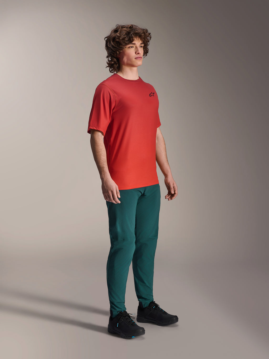 Model draagt Alpinestars A-Dura Pants in Dark Pine, gecombineerd met een rood A-Dura Rise Jersey met korte mouwen en zwarte MTB-schoenen, sportieve mountainbike-kleding in een studio-omgeving