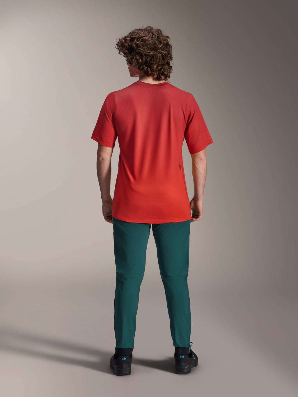 Alpinestars A-Dura Rise Jersey - Korte Mouwen, Spicy Orange, achteraanzicht op een model met een donkergroene broek en zwarte schoenen, voorzien van een verlengde achterkant en een klein logo op de rug, lichtgewicht performance MTB-kleding.