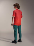 Alpinestars A-Dura Rise Jersey - korte mouwen in Spicy Orange, achteraanzicht van model dat het fietsshirt draagt met een donkergroene mountainbikebroek en zwarte schoenen, voorzien van een verlengde achterzijde en performance-pasvorm voor off-road rijden