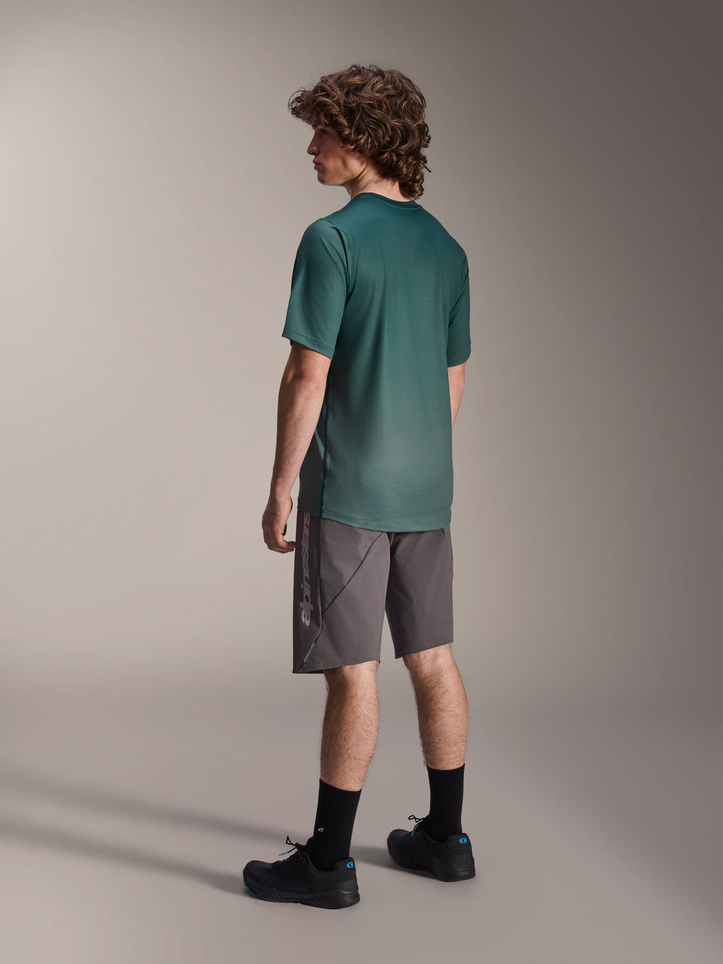 Alpinestars A-Dura Rise Jersey, MTB-shirt met korte mouwen, Silver Pine groen, gedragen met grijze short en zwarte schoenen, achteraanzicht met verlengde achterkant en regular fit, lichtgewicht vochtafvoerende stof voor trailrijden