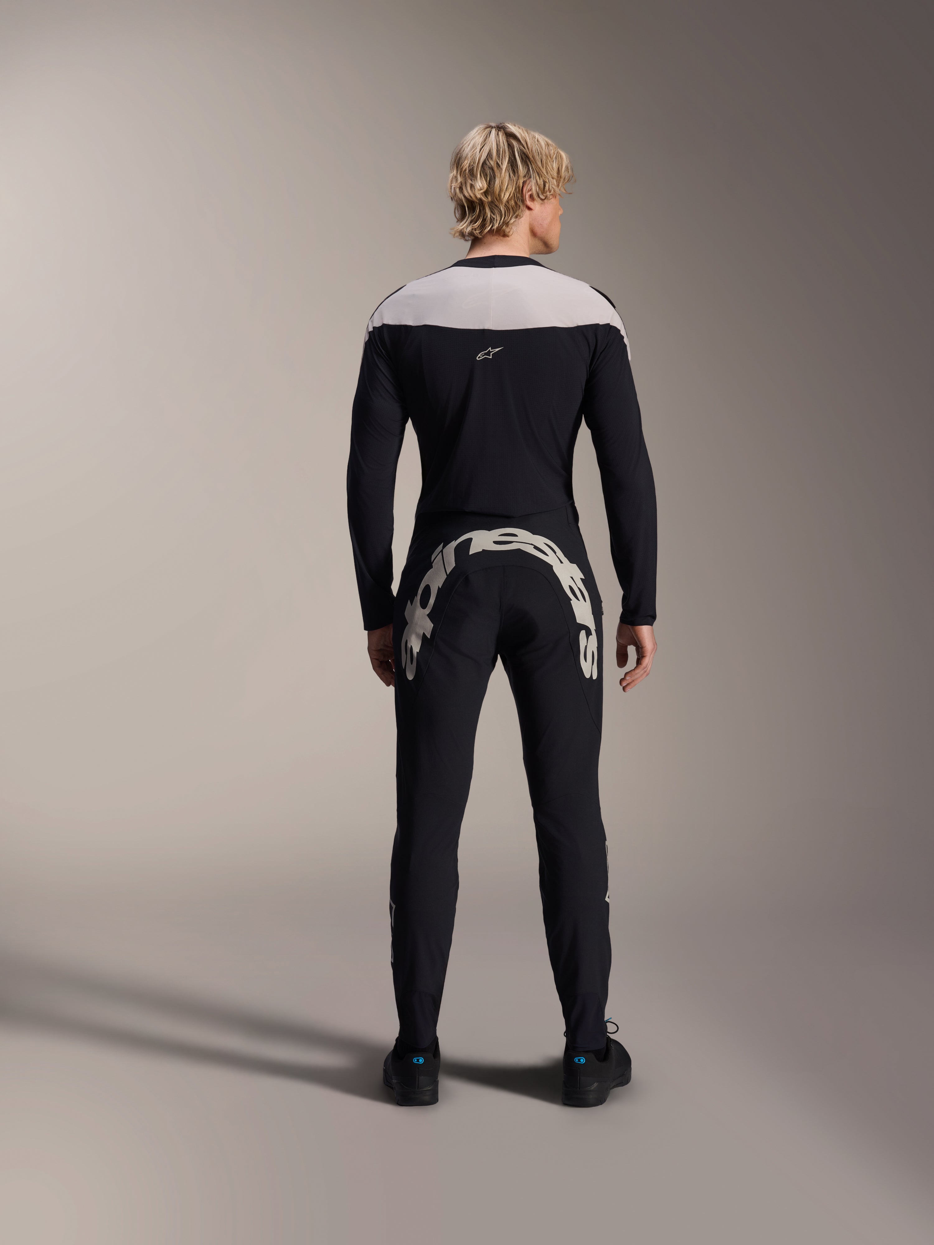 Alpinestars A-Supra Melt jersey met lange mouwen, zwart met lichtgrijs schouderpaneel, zwarte MTB-broek met groot Alpinestars-logo op de achterkant, man van achteren gezien, performance mountainbike-uitrusting