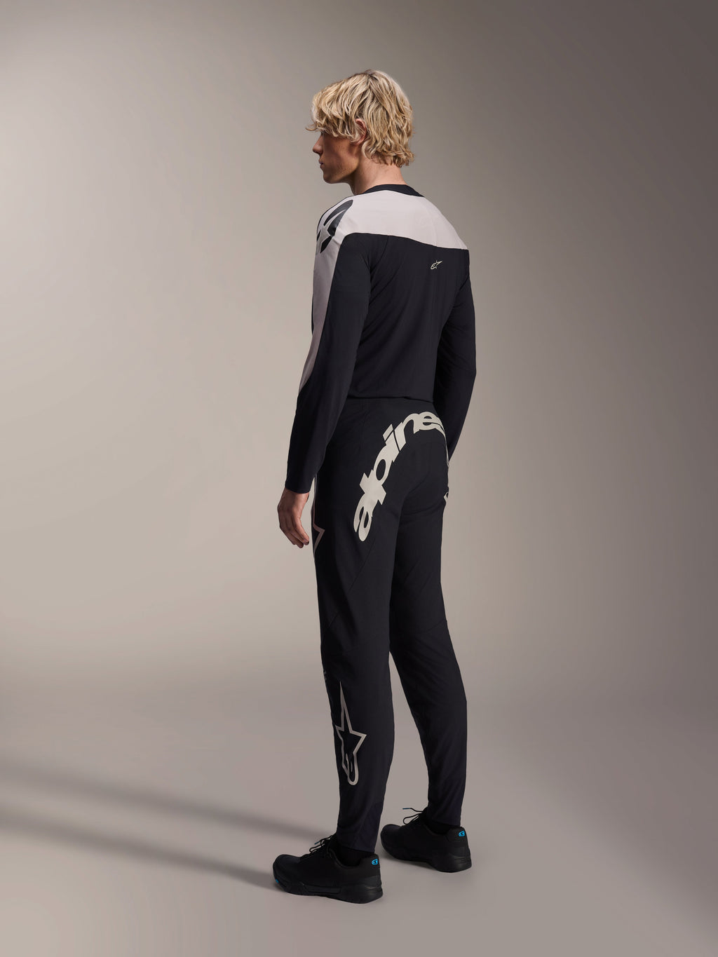 Alpinestars A-Supra Melt broek, MTB-broek, zwart, voorzien van een groot wit Alpinestars-woordmerk op de achterpas en een sterlogo op de broekspijp, gedragen door een model met een bijpassende jersey met lange mouwen en zwarte schoenen, zij-achteraanzicht