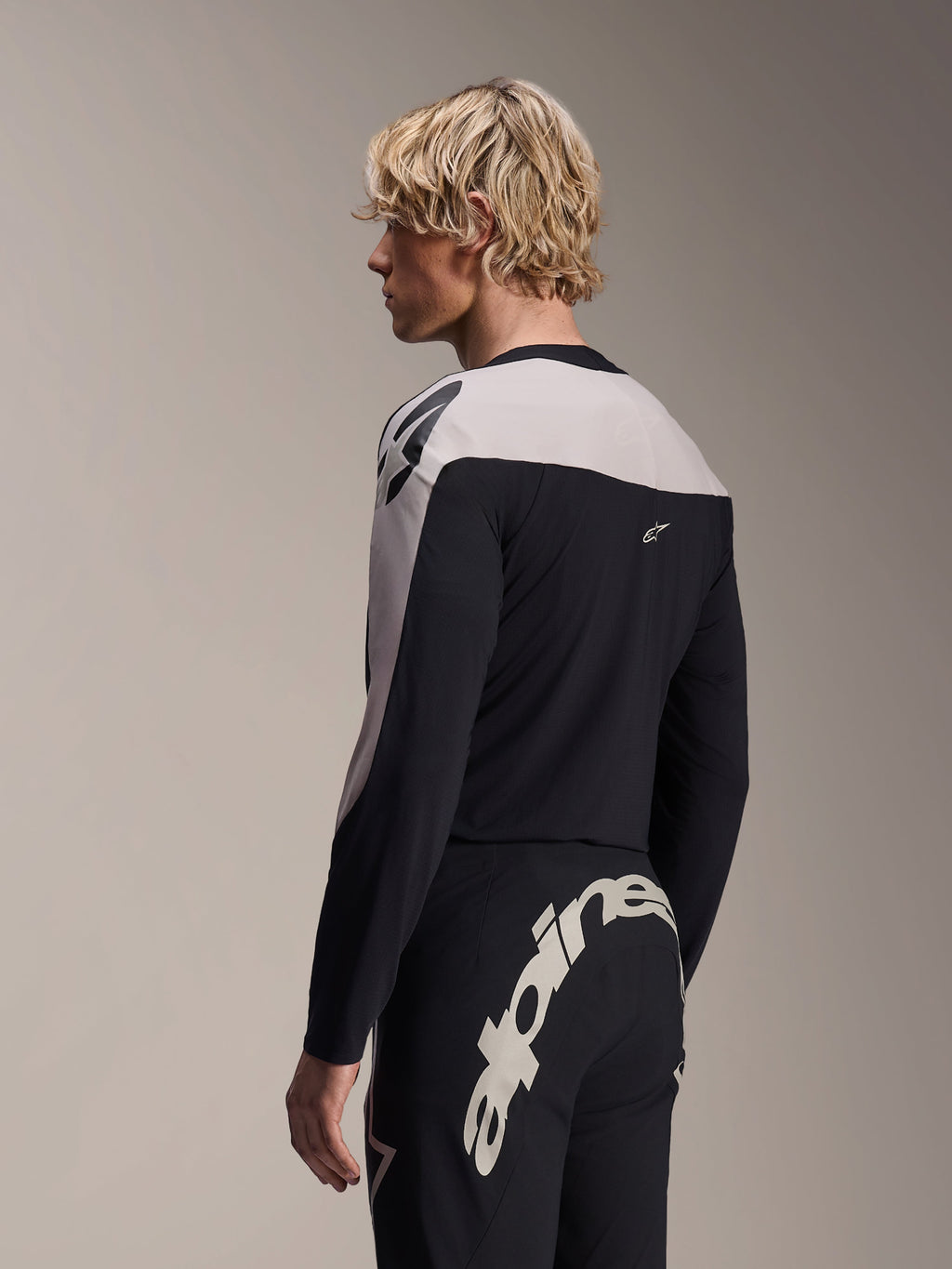 Mannelijk model draagt Alpinestars A-Supra Melt Jersey met lange mouwen en A-Supra Melt broek, zwart en lichtgrijs MTB-racetenue, met groot wit Alpinestars-woordmerk over de achterpas van de broek