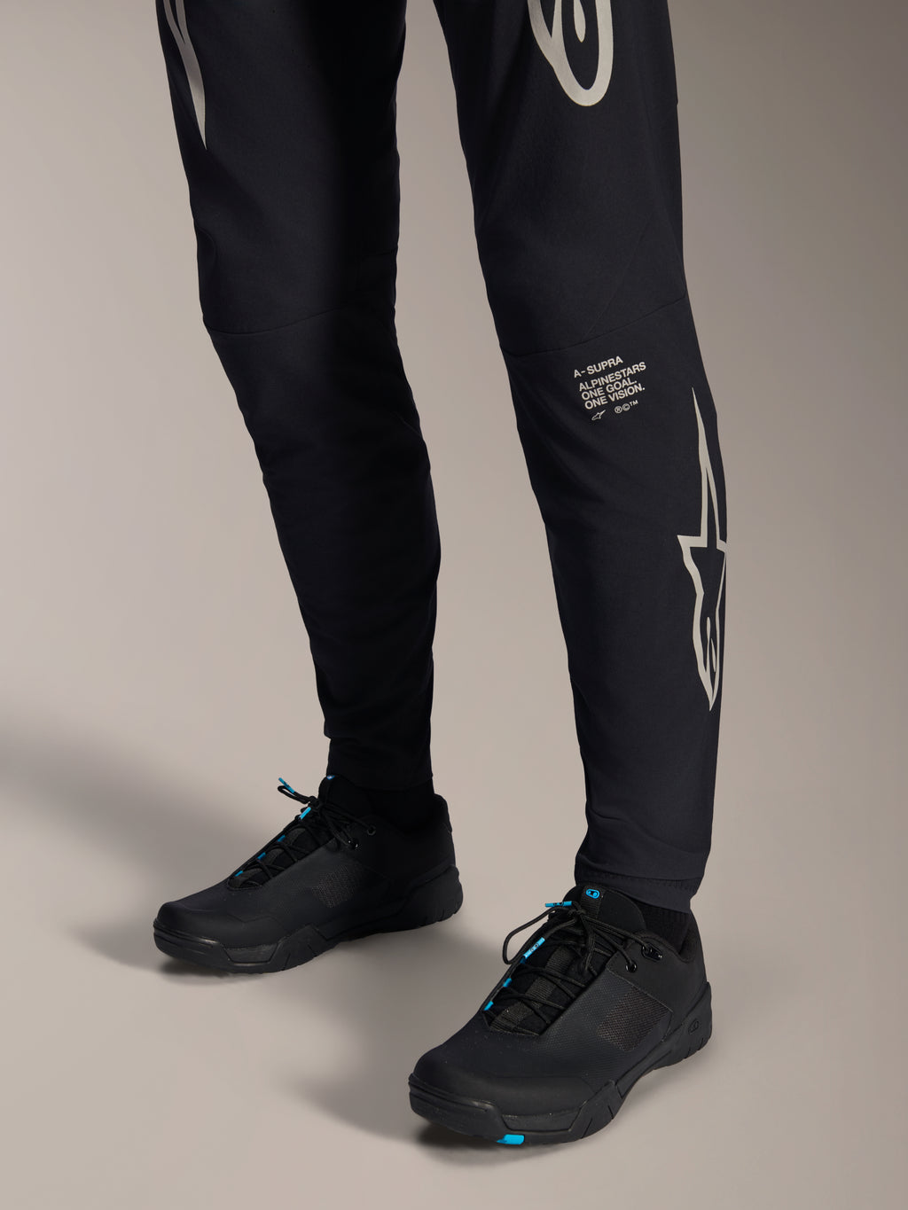 Alpinestars A-Supra Melt Pants, zwarte mountainbike broek, met wit Alpinestars-woordmerk en logo op het been, gedragen met zwarte fietsschoenen met blauwe accenten, performance off-road uitrusting
