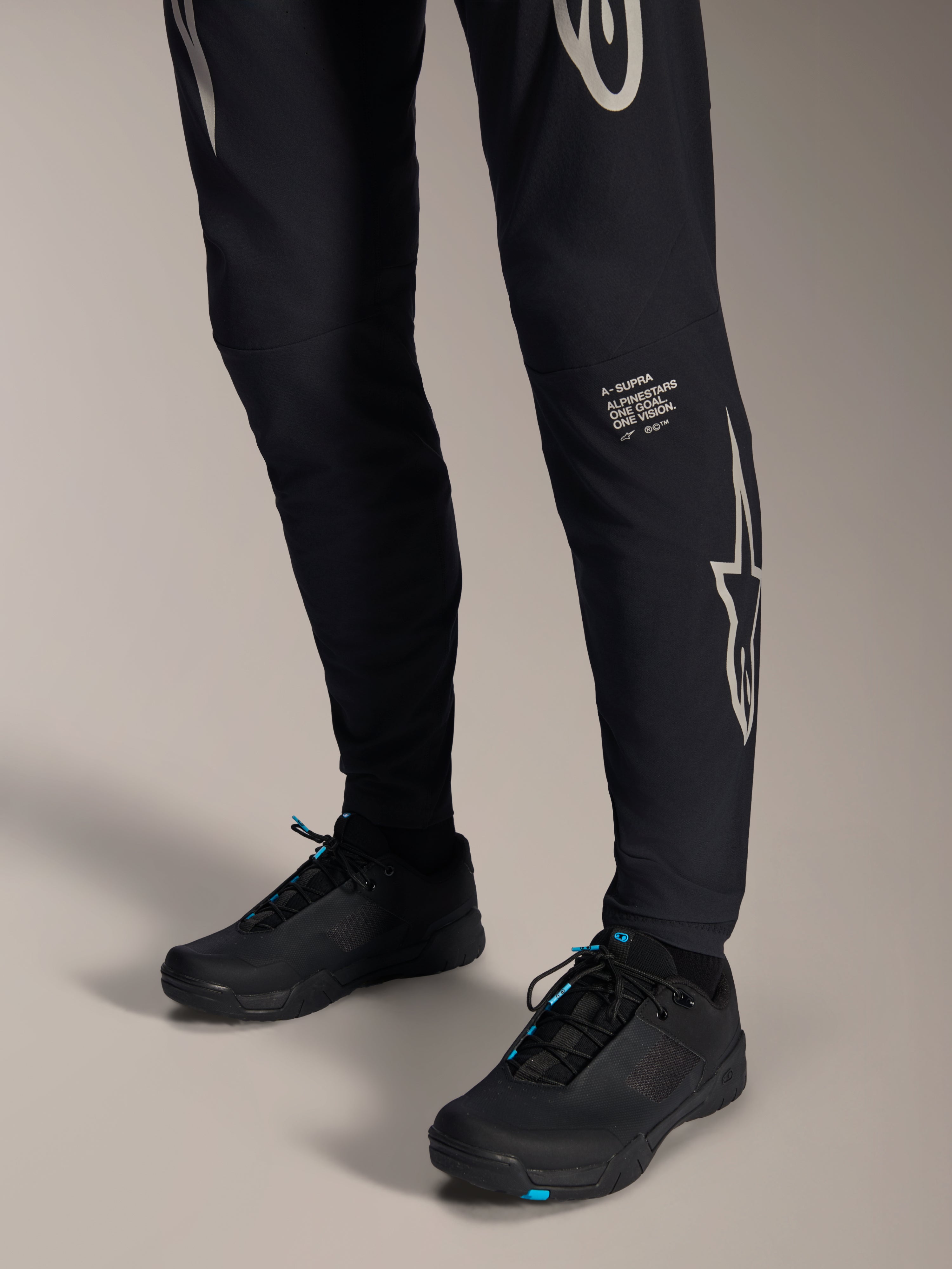 Alpinestars A-Supra Melt Pants, zwarte mountainbike broek, met wit Alpinestars-woordmerk en logo op het been, gedragen met zwarte fietsschoenen met blauwe accenten, performance off-road uitrusting