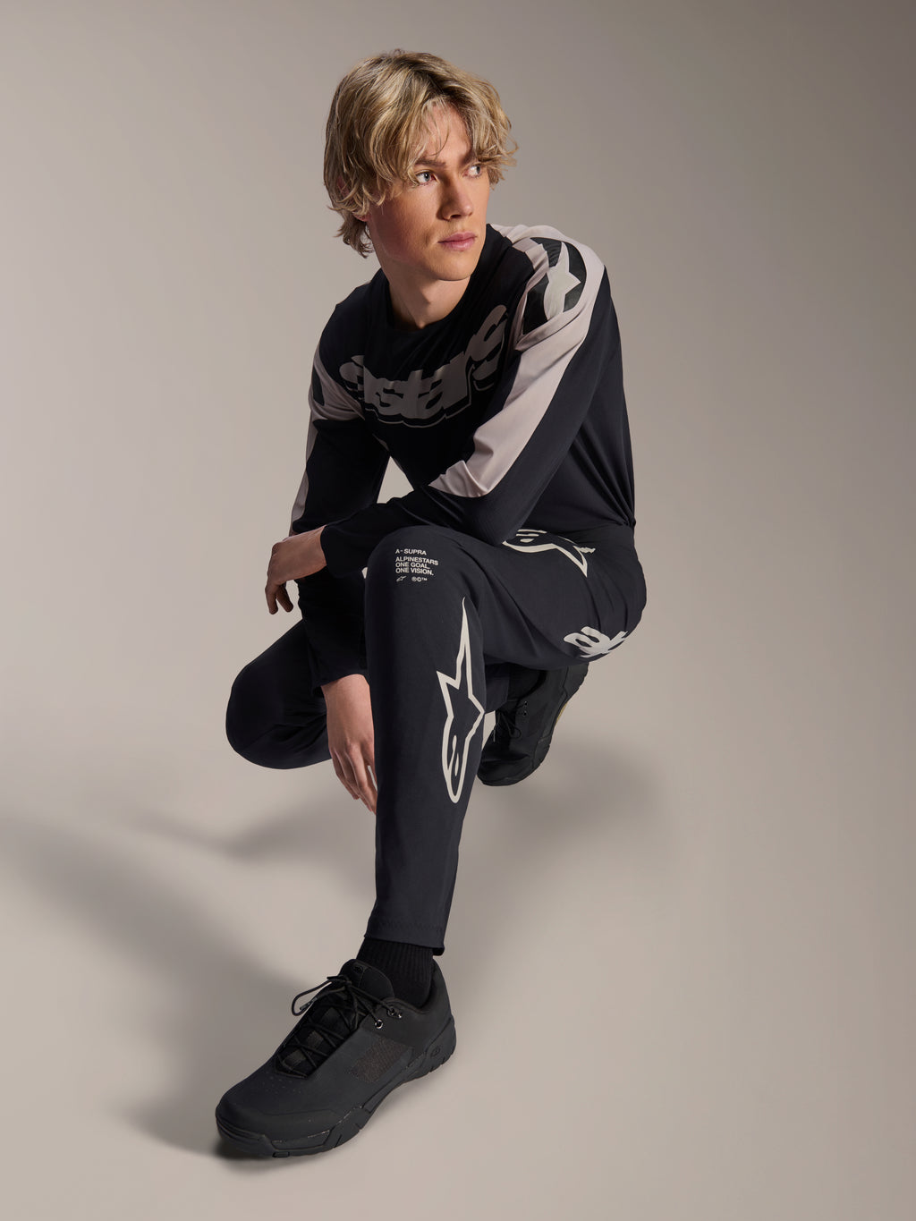 Alpinestars A-Supra Melt broek en jersey met lange mouwen, zwart en grijs, voorzien van opvallende witte branding en sterlogo's, gedragen door een mannelijk model in een gehurkte houding, gecoördineerde MTB-race-uitrusting