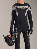 Alpinestars A-Supra Melt MTB-raceset, jersey met lange mouwen en broek, zwart en grijs met witte Astars-logo's, model houdt een zwarte full-face helm vast en draagt bijpassende handschoenen, hoogwaardige uitrusting voor downhill- en enduro-mountainbiken