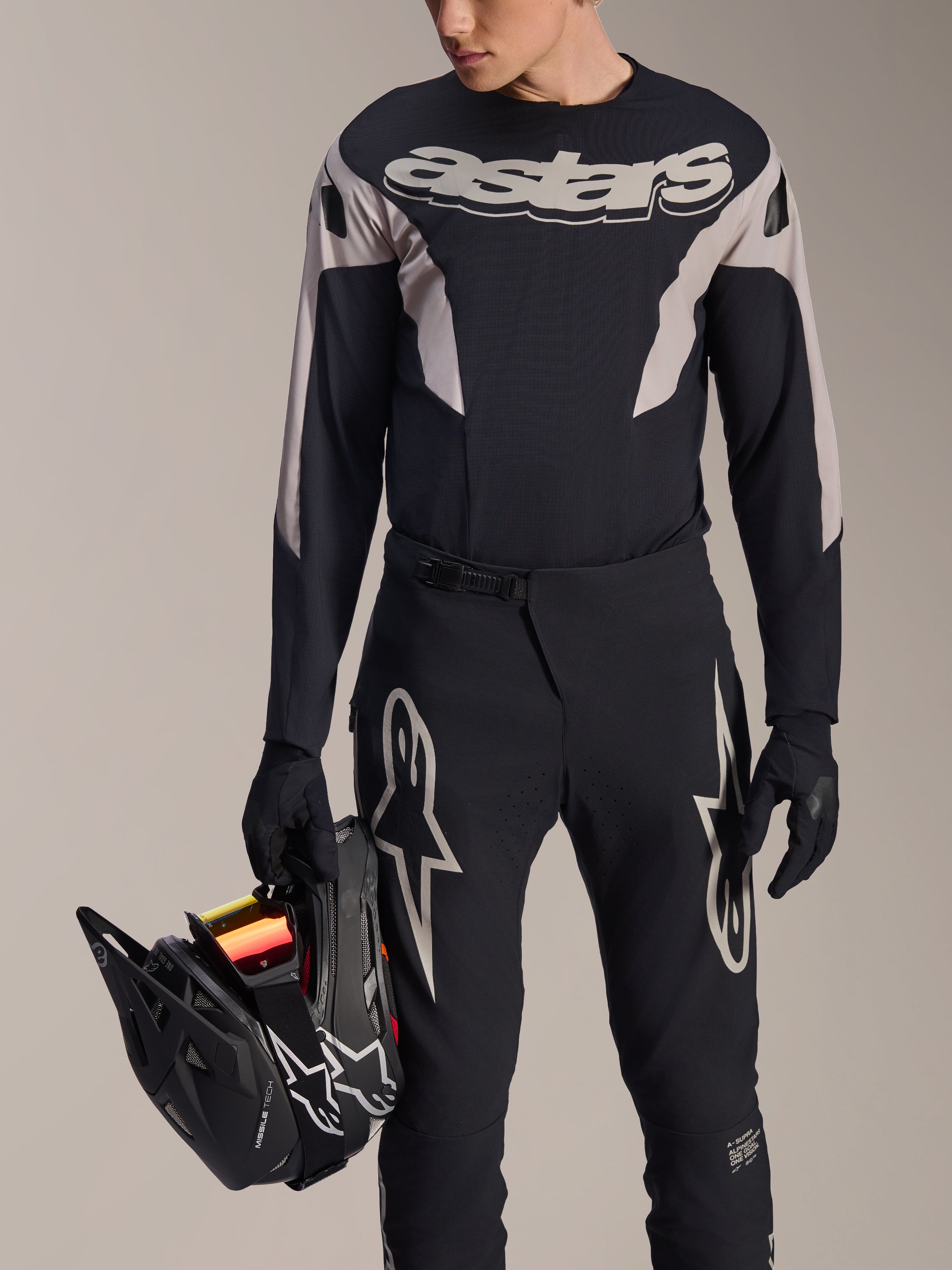 Alpinestars A-Supra Melt jersey met lange mouwen, zwart met grijze grafische panelen en opvallend 'astars' woordmerk op de borst, gedragen met een zwarte mountainbikebroek en handschoenen terwijl een zwarte full-face helm wordt vastgehouden, performance MTB-race-uitrusting