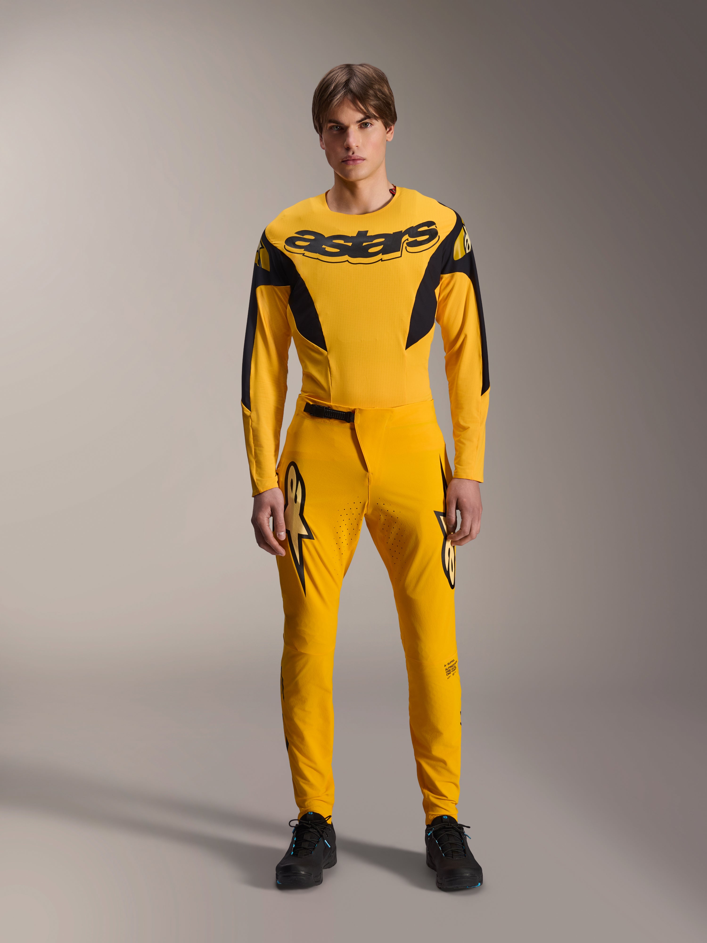 Limited Edition A-Supra Melt Gold-uitrustingsset, Alpinestars mountainbikekleding, donkergeel en zwart, model draagt jersey met lange mouwen met opvallend 'astars'-logo op de borst en bijpassende performance-broek, voorzien van lasergesneden ventilatie op de binnendijen en een veilige ratelsluiting, professionele MTB-racekit