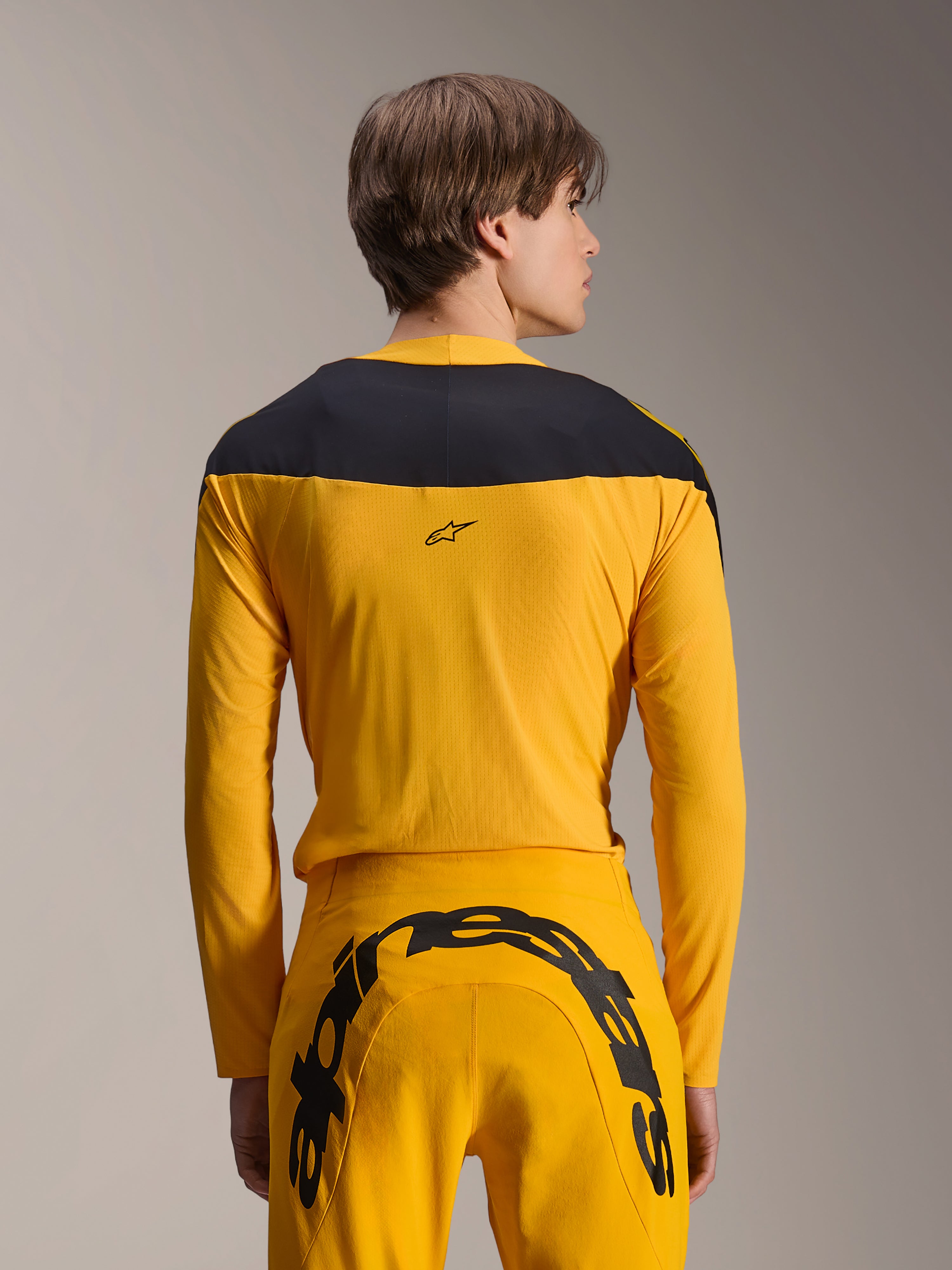 Alpinestars Limited Edition A-Supra Melt Gold Gear-Set, gele MTB-kledingset, achteraanzicht van model met jersey met lange mouwen en zwart schouderpaneel en bijpassende gele broek met groot zwart logo op het achterpand, hoogwaardige fietskleding.