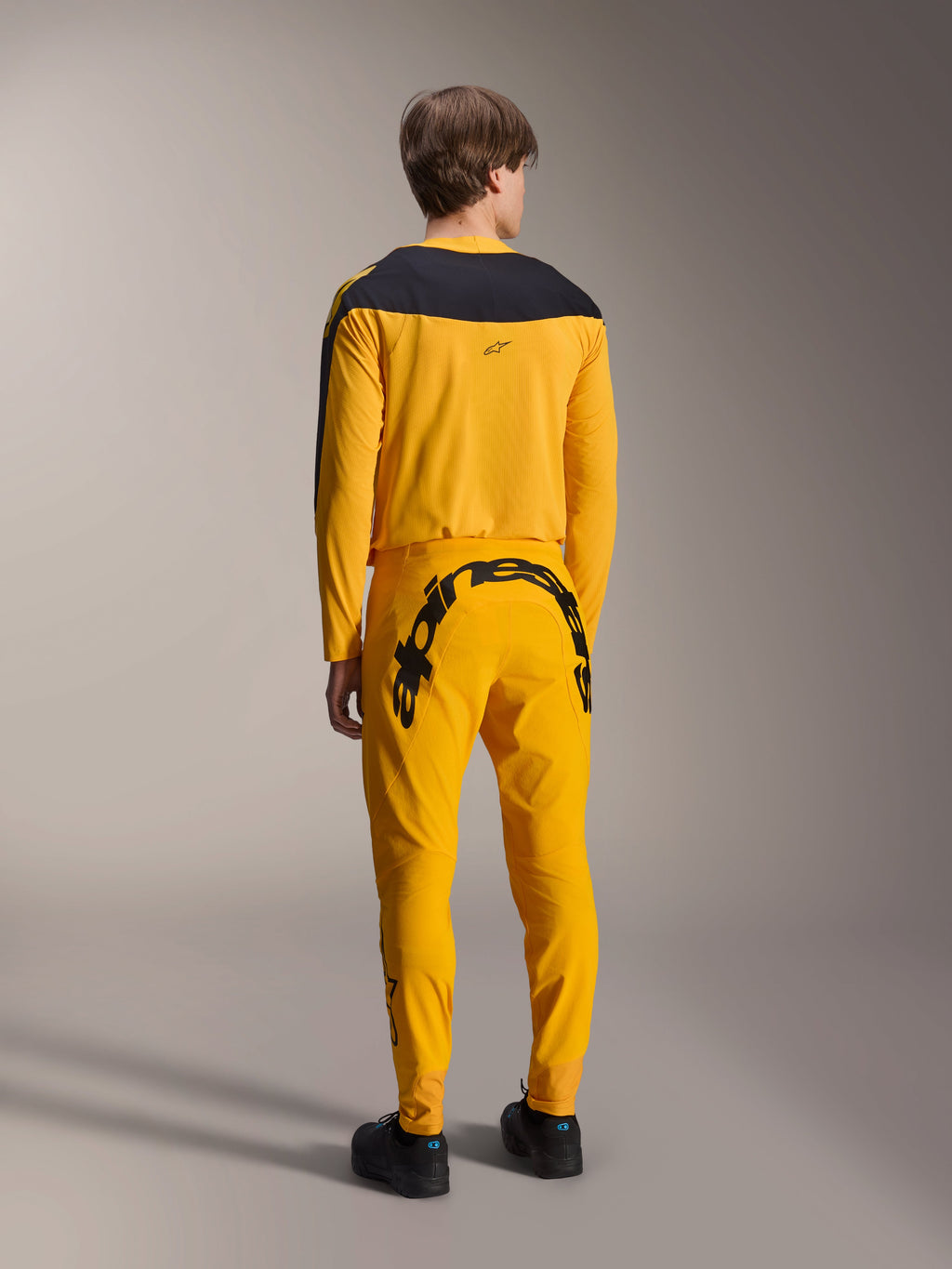 Alpinestars Limited Edition A-Supra Melt Gold Jersey, MTB-jersey met lange mouwen, donkergeel met zwart schouderpaneel, gedragen met bijpassende gele broek met opvallende zwarte branding op de achterzijde, achteraanzicht van prestatiegerichte fietsuitrusting