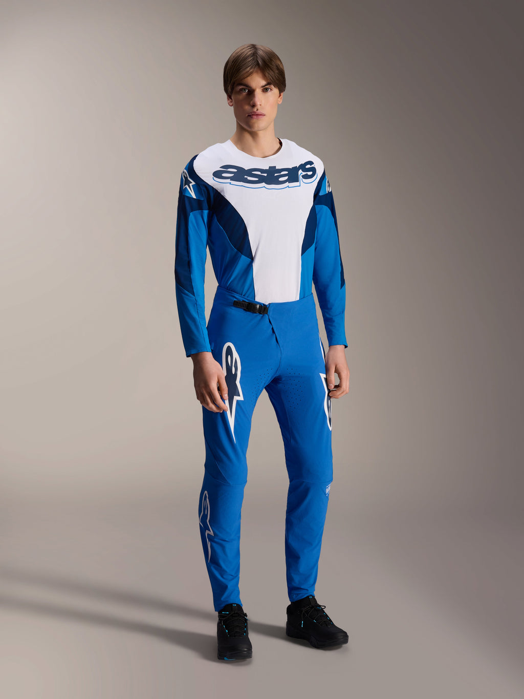 Mannelijk model in de Alpinestars A-Supra Melt Jersey met lange mouwen in blauw en wit, voorzien van een opvallend 'astars'-logo op de borst, gecombineerd met een bijpassende blauwe mountainbikebroek en zwarte schoenen, prestatiegerichte MTB-racekleding.