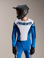 Model draagt Alpinestars A-Supra Melt Jersey met lange mouwen in blauw en wit, voorzien van 'astars' woordmerk op de borst, gecombineerd met bijpassende blauwe MTB-broek, zwarte full-face helm en goggle, complete mountainbike race-outfit.
