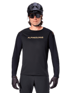Alpinestars A-Aria Polartec Switch Jersey met lange mouwen, zwart, goudkleurig Alpinestars-logo op de borst, gedragen met een zwarte MTB-helm, donkere zonnebril en zwarte handschoenen, professionele mountainbike trailuitrusting