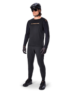 Alpinestars A-Aria Polartec Switch jersey met lange mouwen, zwart, bronskleurige branding op de borst, gedragen door mountainbiker met zwarte helm en zonnebril, hoogwaardige fietskleding voor trailrijden