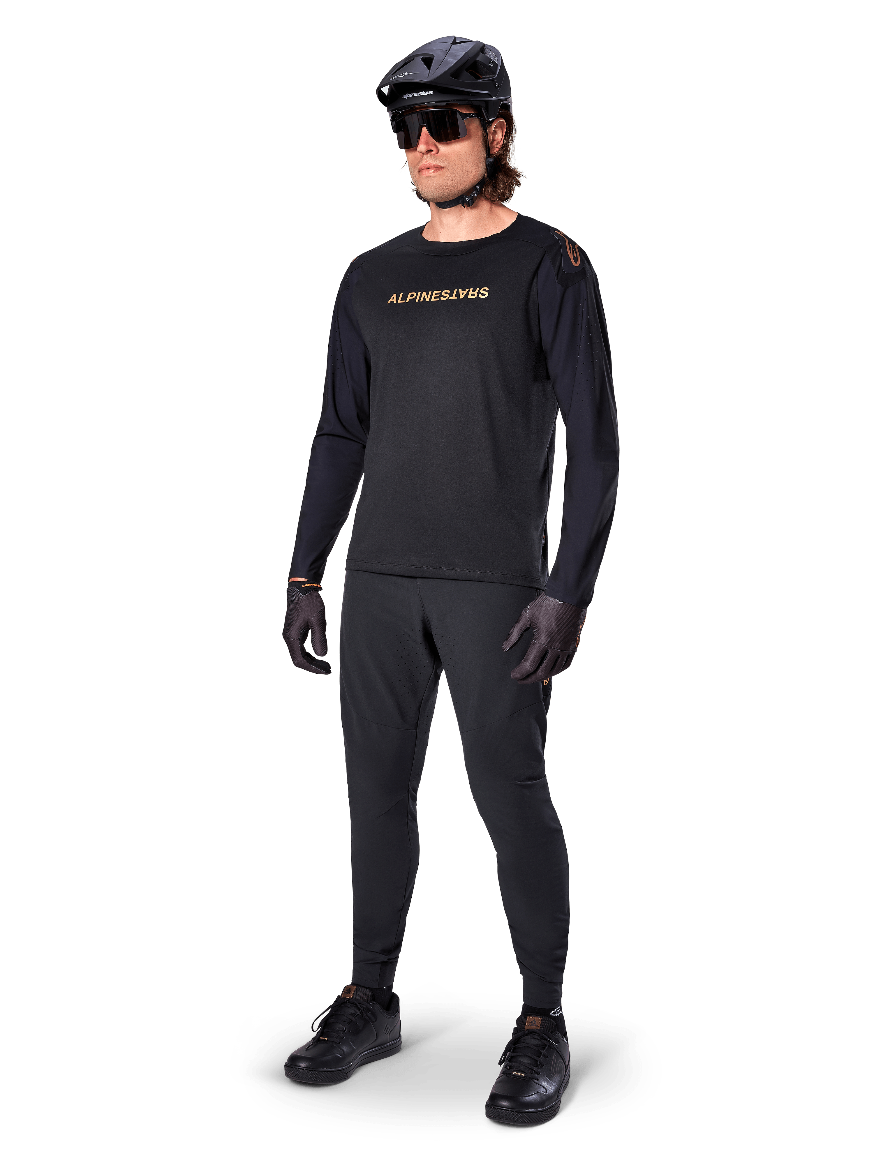 Alpinestars A-Aria Polartec Switch jersey met lange mouwen, zwart, bronskleurige branding op de borst, gedragen door mountainbiker met zwarte helm en zonnebril, hoogwaardige fietskleding voor trailrijden