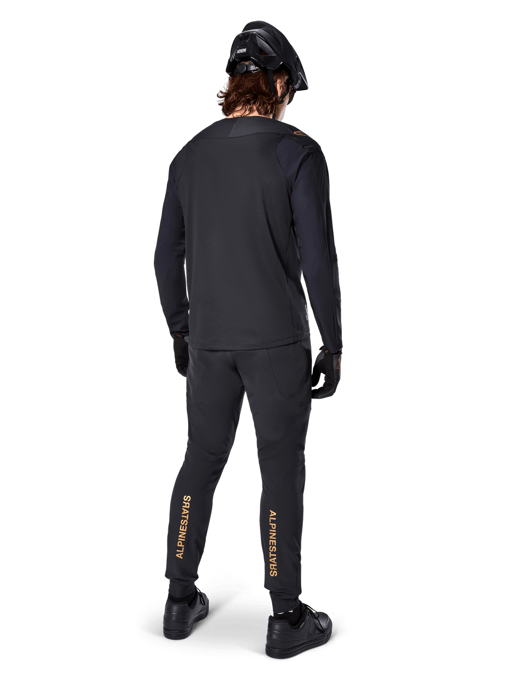 Achteraanzicht van een mountainbiker die het zwarte Alpinestars A-Aria Polartec Switch Jersey met lange mouwen draagt, gecombineerd met een zwarte MTB-broek met lichtbruine Alpinestars-branding op de kuiten, zwarte helm en handschoenen, professionele wieleruitrusting voor off-road paden